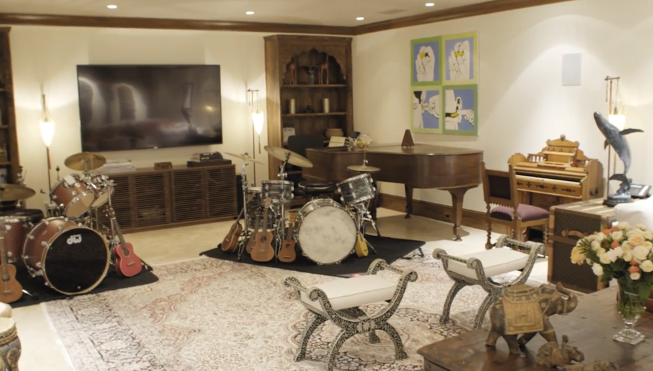 A spacious music room and lounge features a grand piano, multiple drum sets, acoustic guitars, and a vintage pump organ set across a large patterned area rug. Vibrant modern art, carved wooden shelving, and comfortable inlaid benches complete this warm, creative space. | Source: YouTube/Architectural Digest