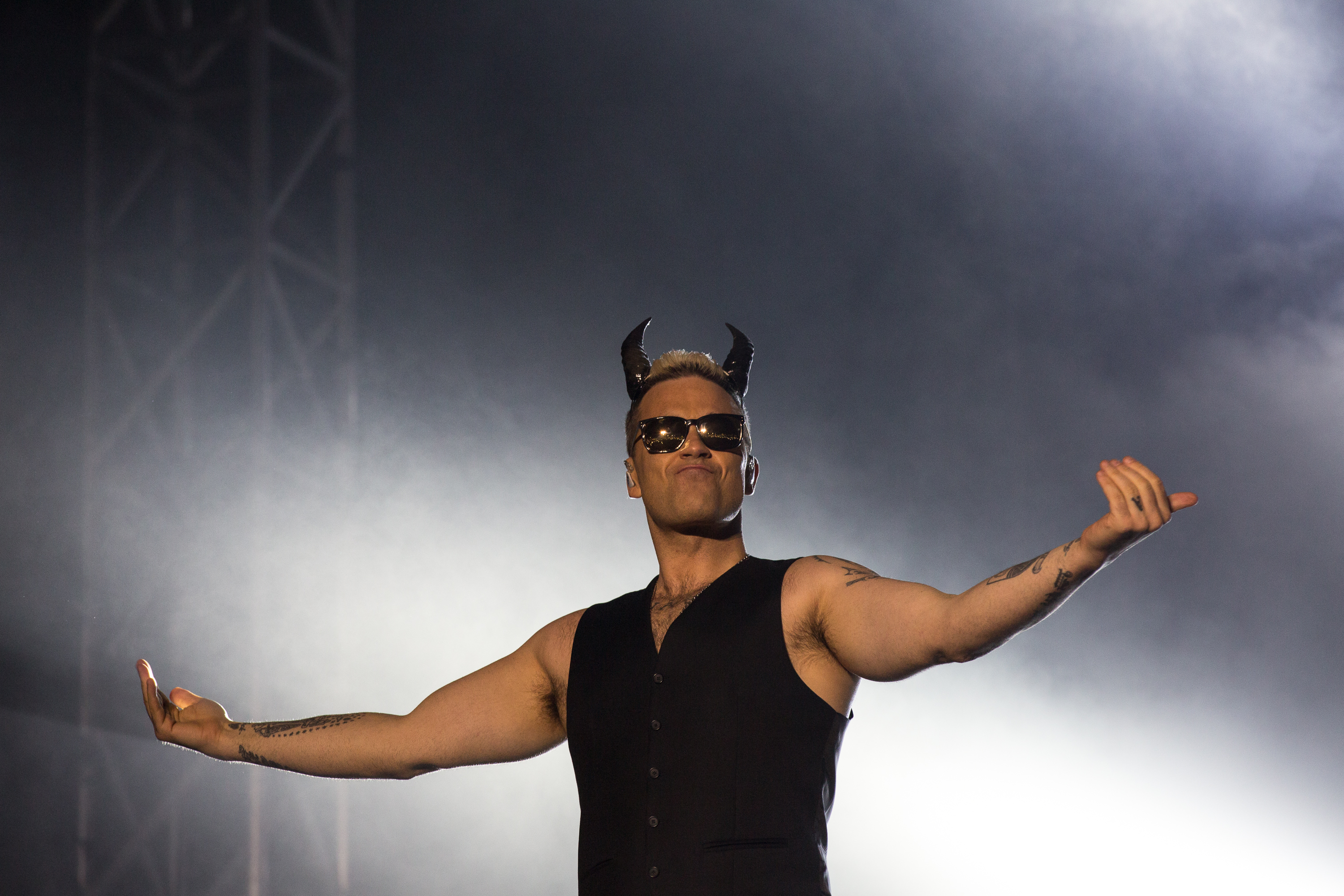 Robbie Williams performed at Hayarkon Park on May 2, 2015, in Tel Aviv, Israel. Wearing sunglasses, a sleeveless vest, and devil horns, he struck a theatrical pose that captured his flair for showmanship and tongue-in-cheek stage persona.