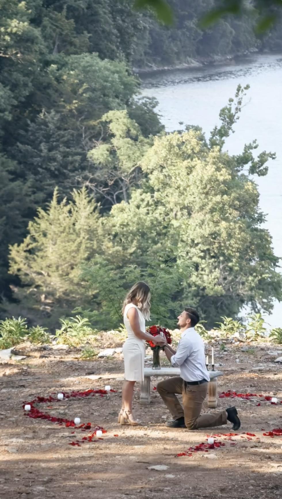 Stephen Wissmann kneels before Jana Duggar, holding a bouquet as he proposes, surrounded by rose petals and candles with the lake in the background. | Source: Instagram/janamduggar