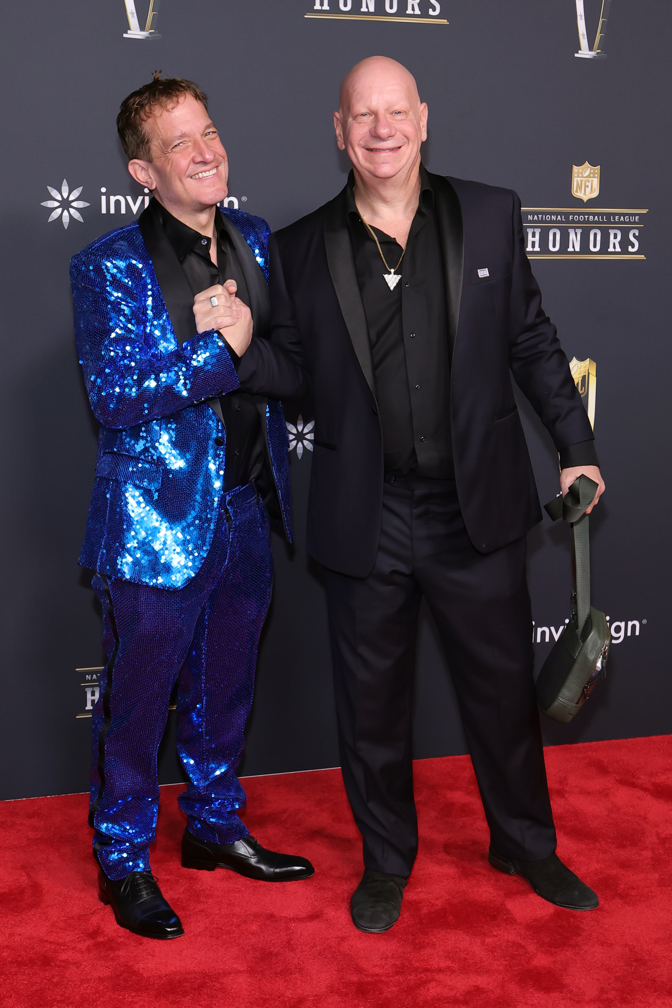 Jeffrey Beacher and Jeff Ross clasp hands and grin, with Jeffrey in a shimmering blue suit and Jeff in black, sharing a lighthearted moment amid the formal festivities.