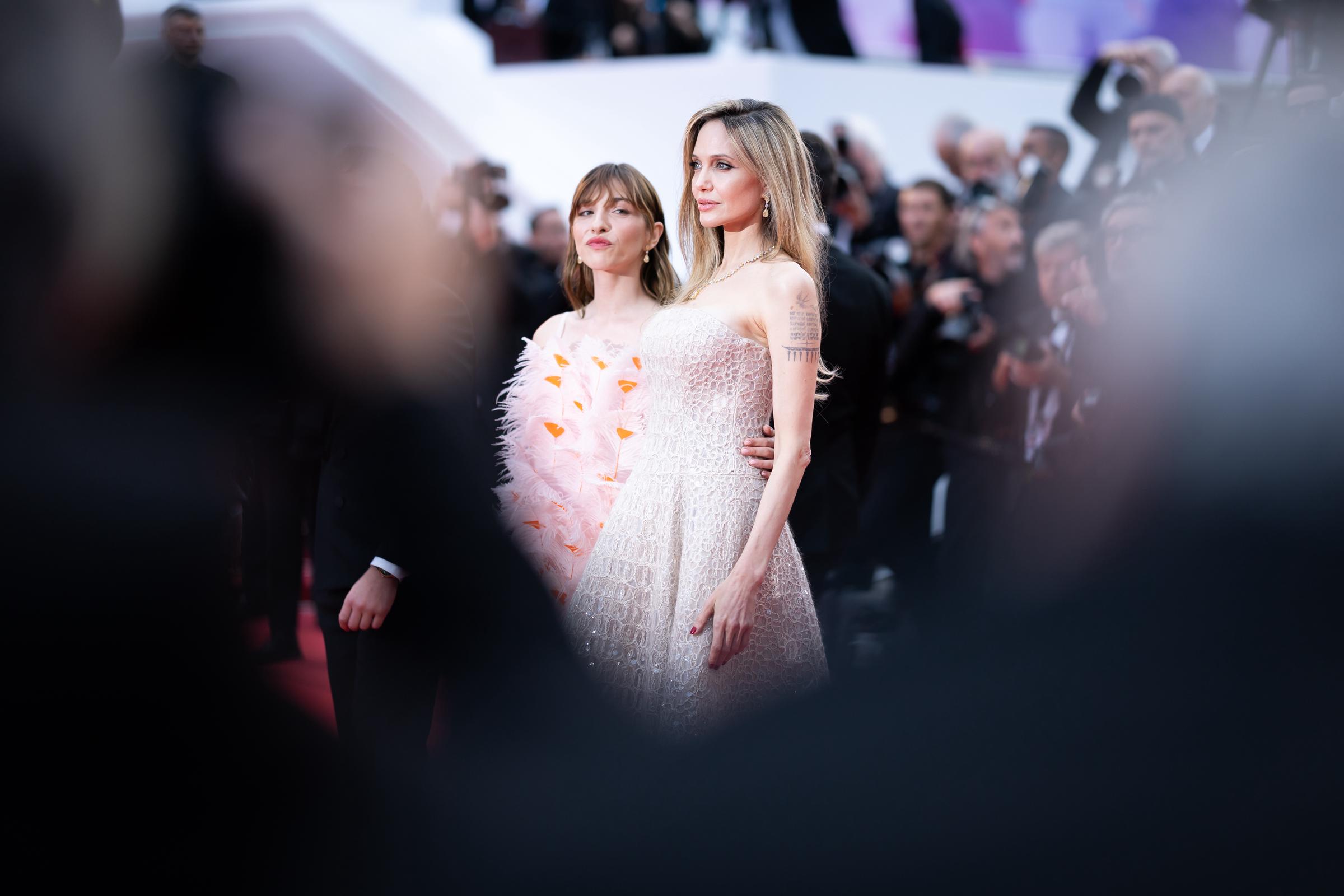 Framed by blurred silhouettes in the foreground, Angelina Jolie and Marie Colomb share a striking moment on the red carpet. Jolie stands tall in her sequined Cucinelli gown, while Colomb stuns in a feathered, floral-inspired mini dress, both women commanding attention from every angle.