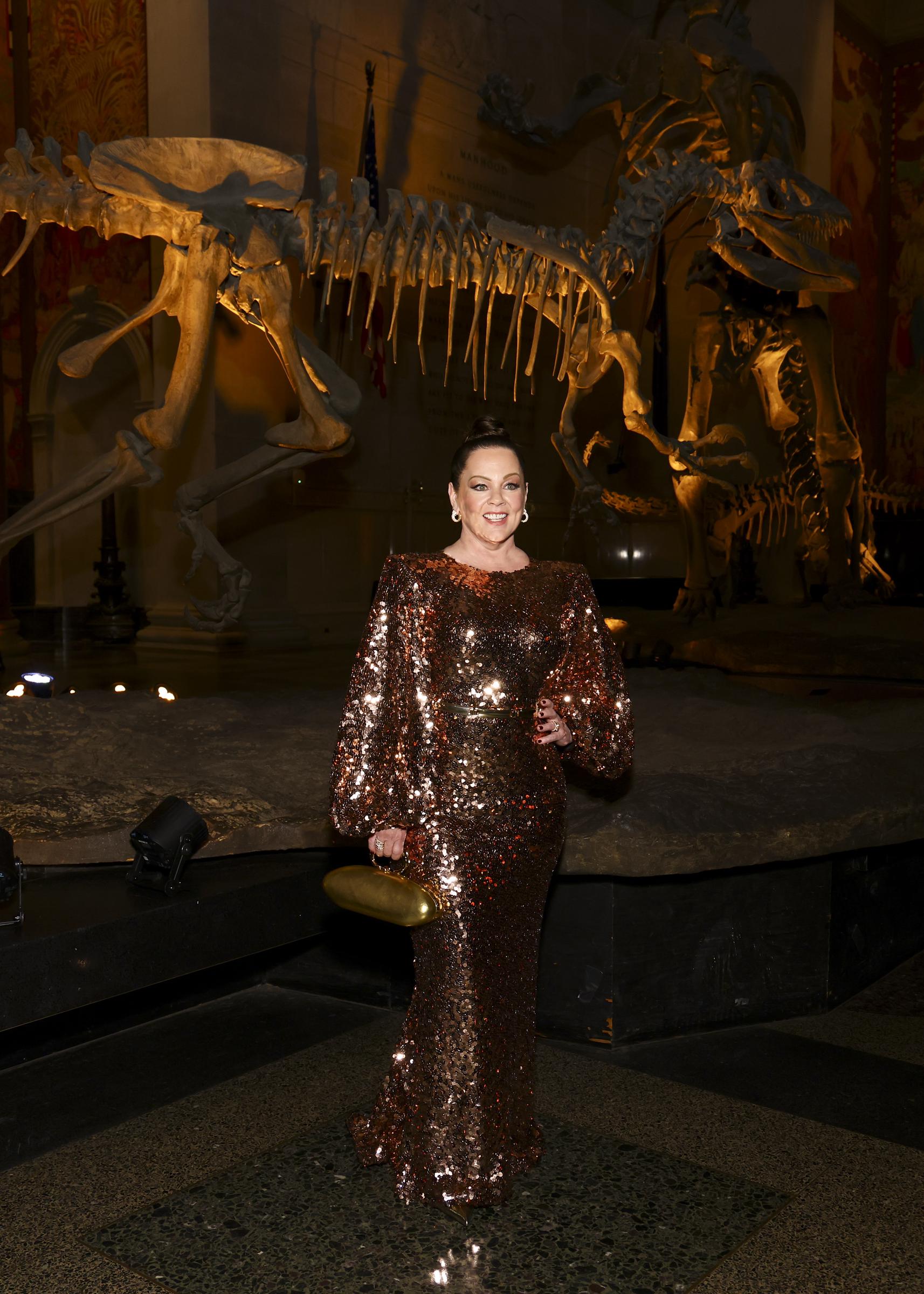 Melisa McCarthy glows in a bronze sequin gown that catches the museum's warm lighting, standing confidently before towering dinosaur fossils. With a golden clutch in hand and a poised smile, she brings modern glamor to a setting shaped by ancient history.