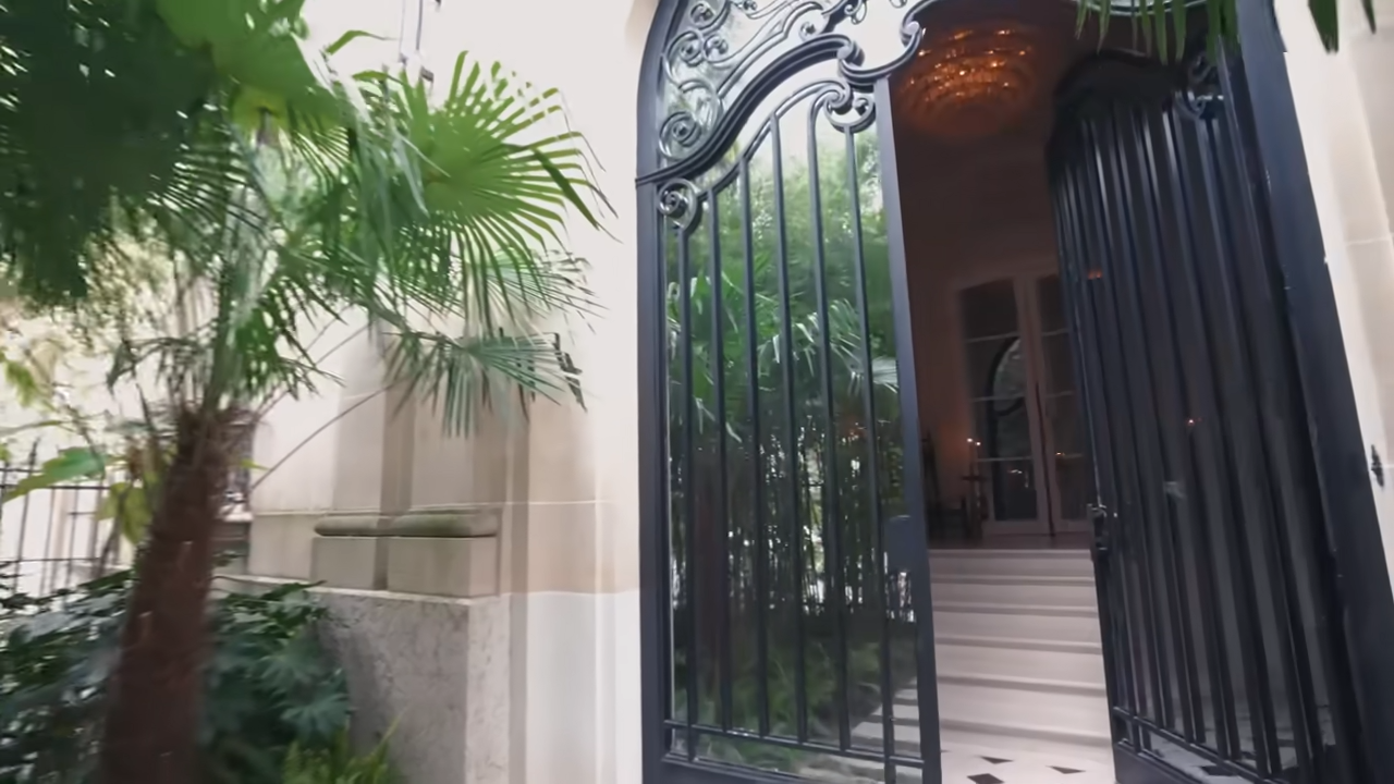 The grand, towering black wrought-iron gates at the entrance of Lenny Kravitz's 1920s Paris townhouse, framed by lush green palms. | Source: YouTube/@Archdigest