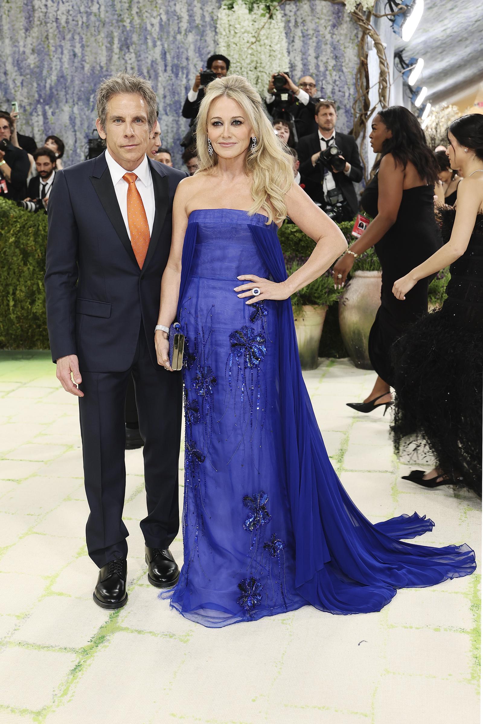 Ben Stiller keeps it classic in a navy suit with a bright orange tie. The clean, tailored look adds a subtle contrast to the night’s bold fashion. Next to him, Christine Taylor shines in a strapless blue gown with floral details and a flowing train.