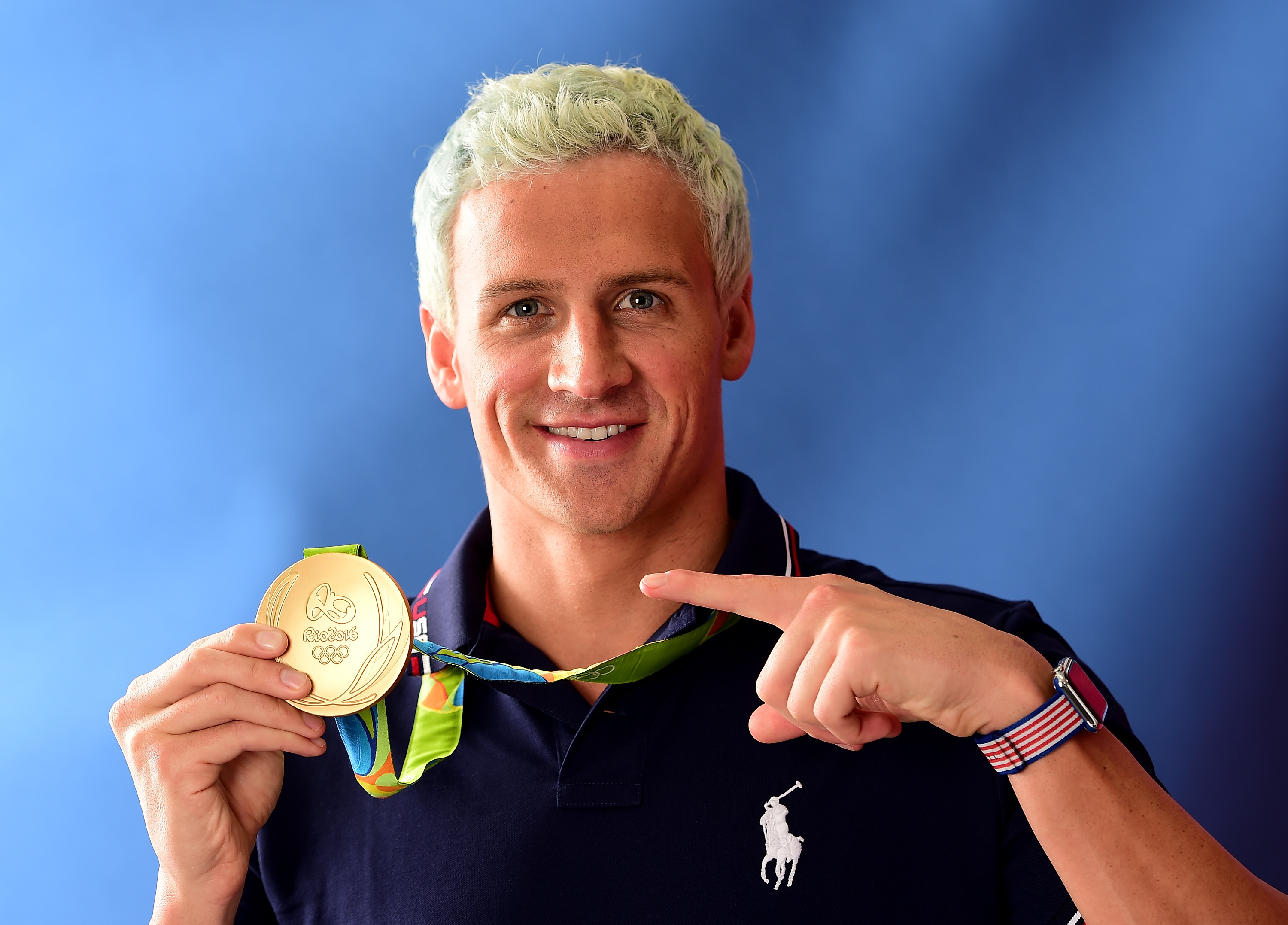 Ryan Lochte proudly points toward his Olympic gold medal while posing on the "Today" show set along Rio’s famous Copacabana Beach. Wearing a navy polo shirt, the swimmer beams as he celebrates another major career achievement.