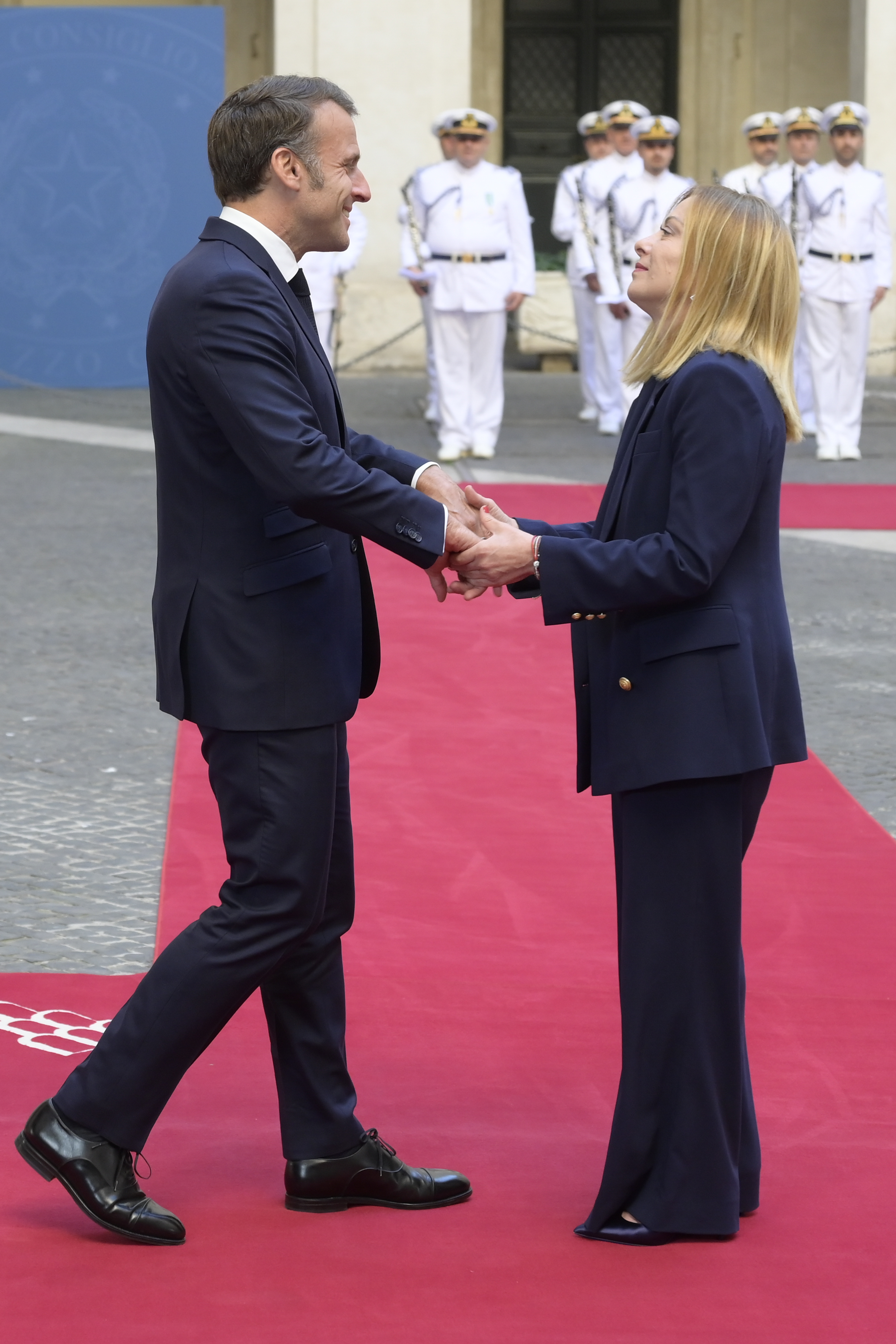 Emmanuel Macron and Giorgia Meloni share a firm handshake while standing on a red carpet outside Palazzo Chigi. Macron flashes a broad smile as Meloni looks up at him, the two surrounded by ceremonial guards in white uniforms.