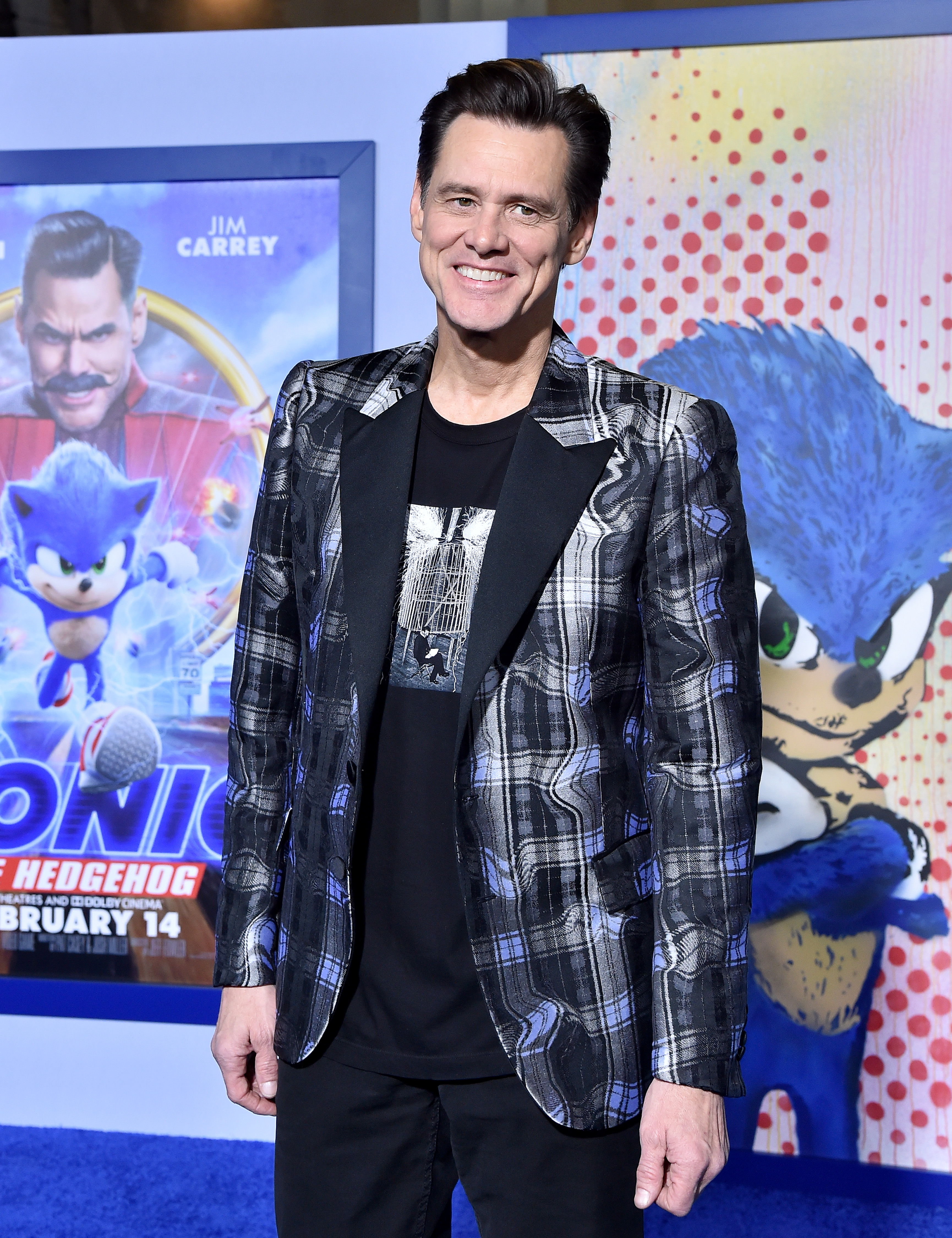 Wearing a patterned plaid blazer over a graphic black tee, Jim Carrey flashes a relaxed smile on the blue carpet. Behind him, vibrant posters of "Sonic the Hedgehog" add a playful pop of color to the screening backdrop.