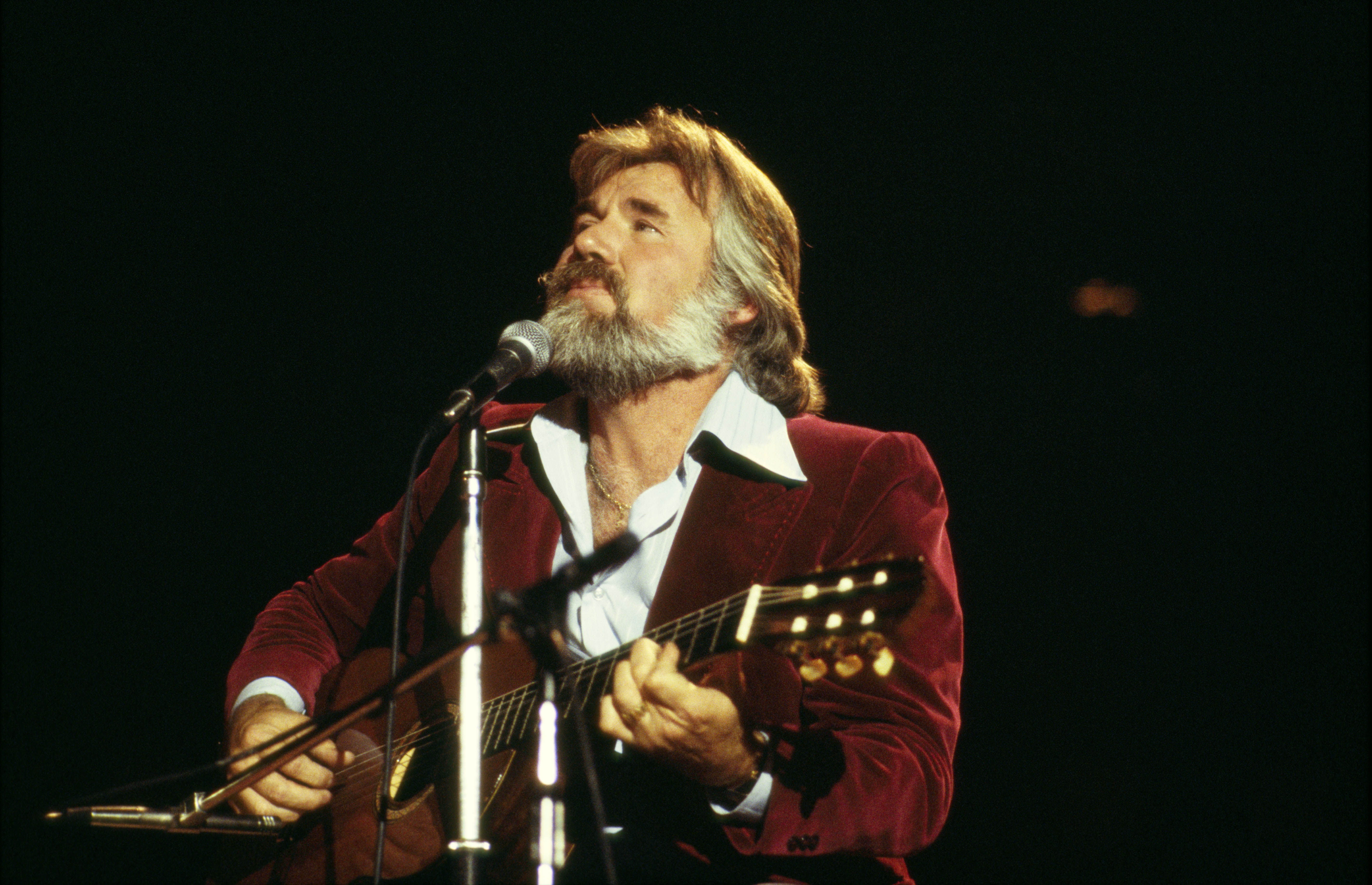 Kenny Rogers performing on stage while playing the guitar wearing a velvet blazer paired with a shirt in 1978. / Source: Getty Images