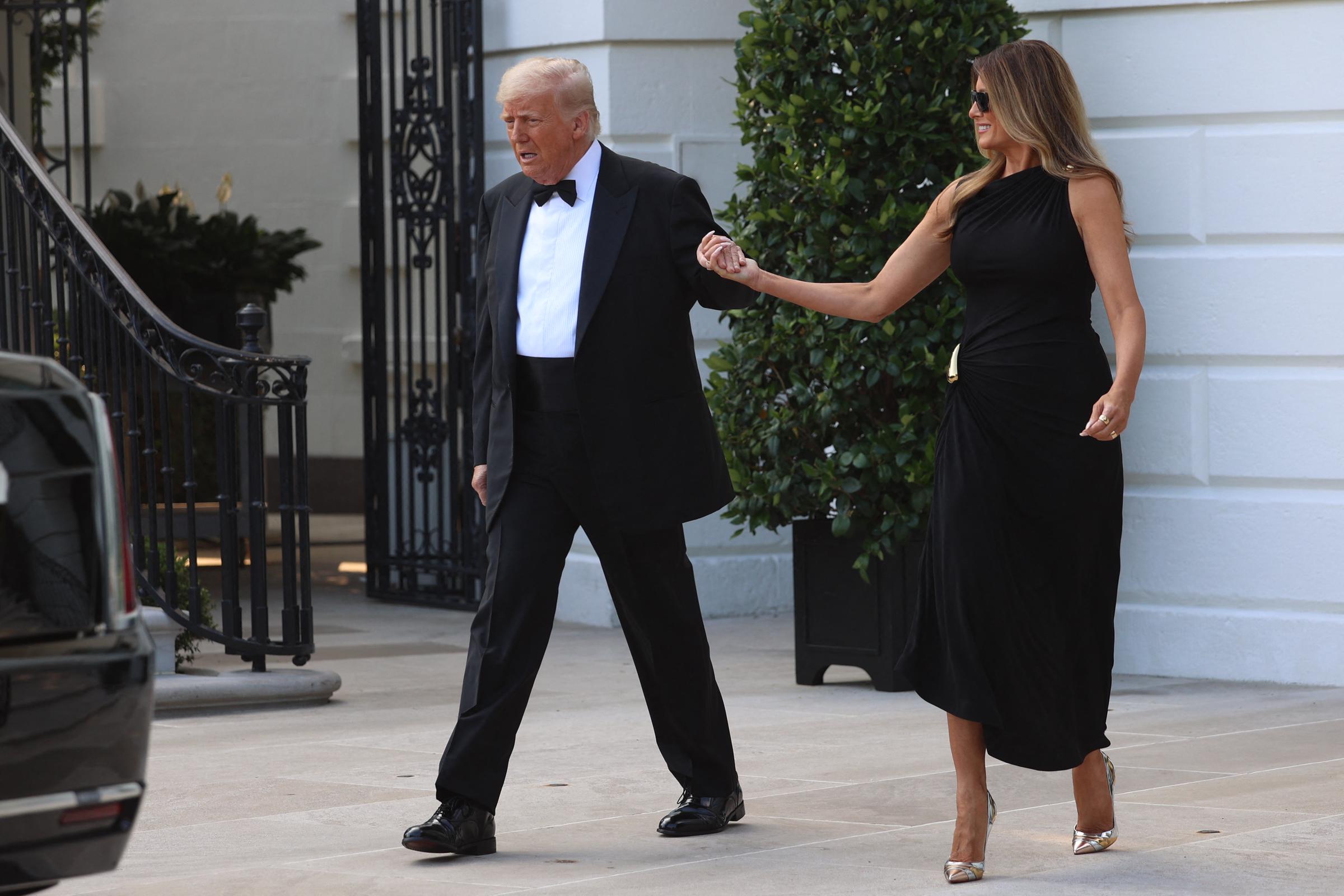 Melania’s entire ensemble is on display as she and her husband leave the White House—her sleek black gown hugs her figure with soft draping at the waist, accented by a gold clasp. She pairs it with metallic pointed-toe heels that catch the light, adding a touch of glamour. Her confident stride and easy smile complete the polished look, while Donald steps forward beside her, hand in hand.