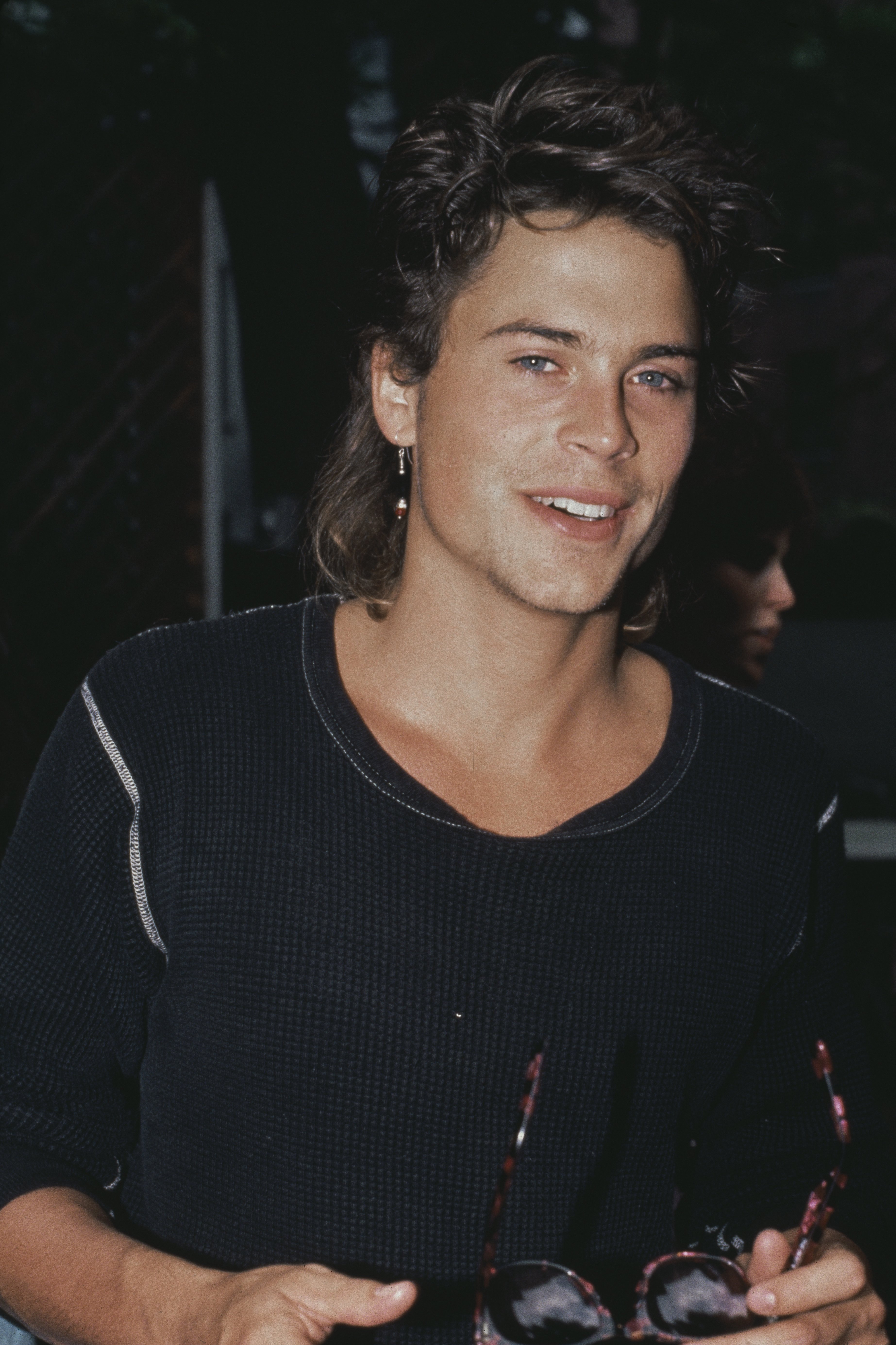 Rob Lowe captured beaming, wearing a black top while holding a pair of sunglasses in 1985. / Source: Getty Images