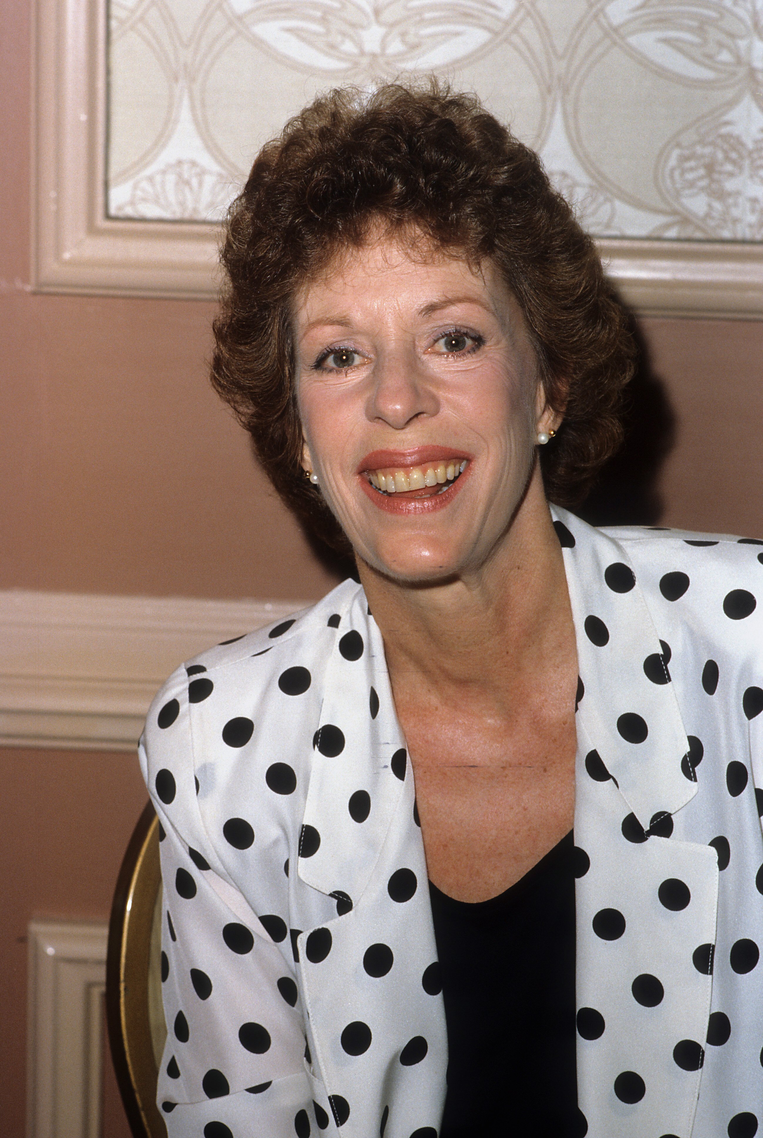 Carol Burnett faces the camera in a close portrait, wearing a white blazer patterned with large black polka dots over a dark top. She is seated, leaning slightly forward, with a direct smile.