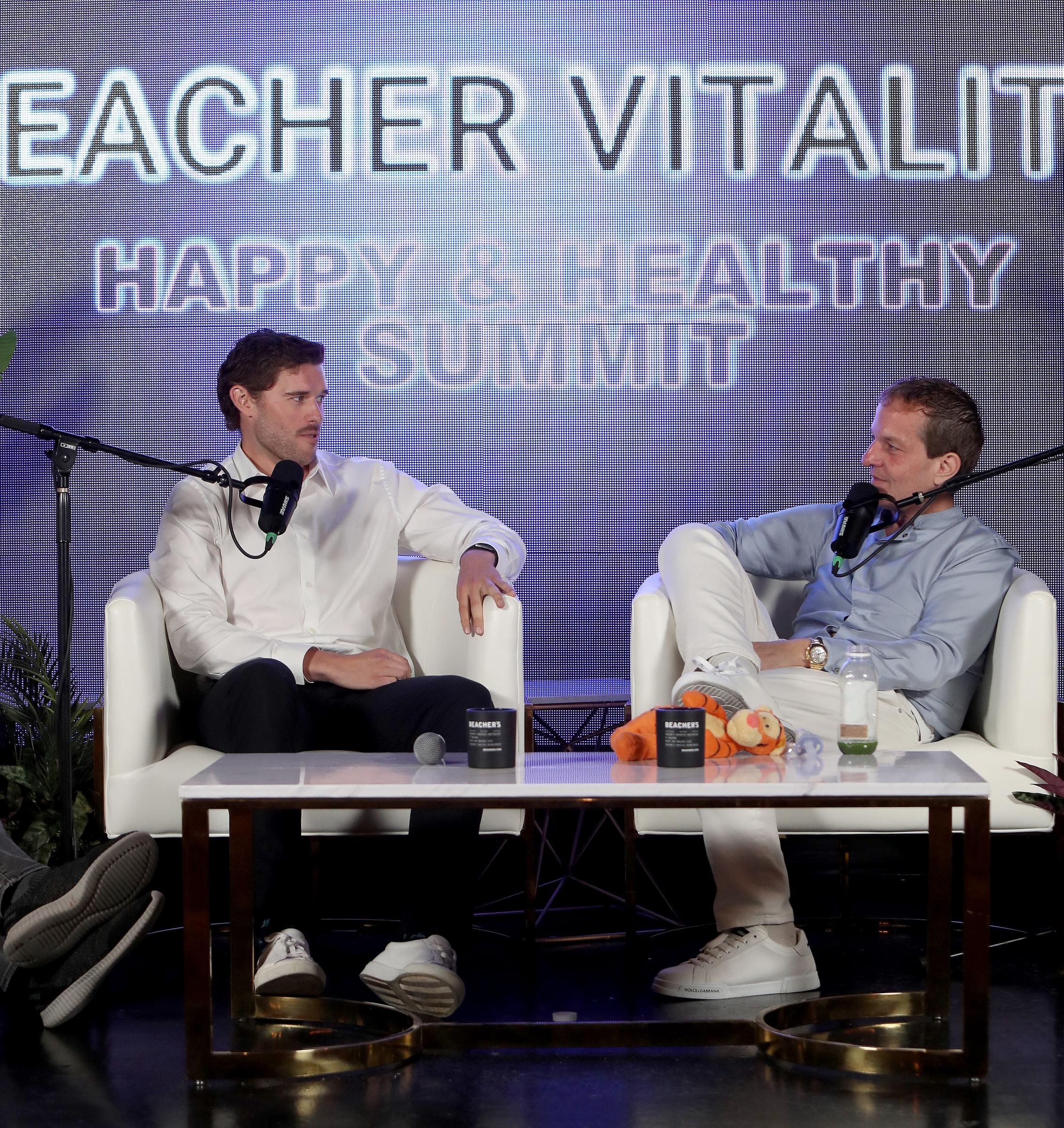 In this wide shot from the summit, Christopher Schwarzenegger maintains a poised demeanor while engaging with Jeff Beacher. The branded backdrop reinforces the event's central message of wellness and transformation.