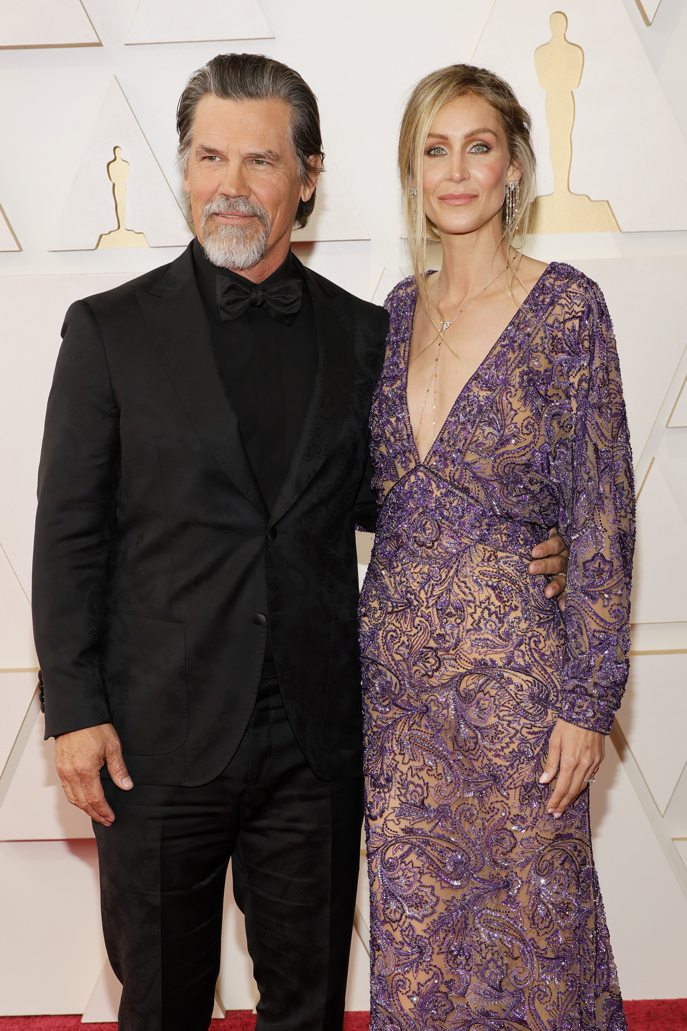 Dressed in an all-black suit with a tonal pattern and matching bow tie, he appears with a full salt-and-pepper beard and slicked-back hair. His look is mature, composed, and carefully styled.