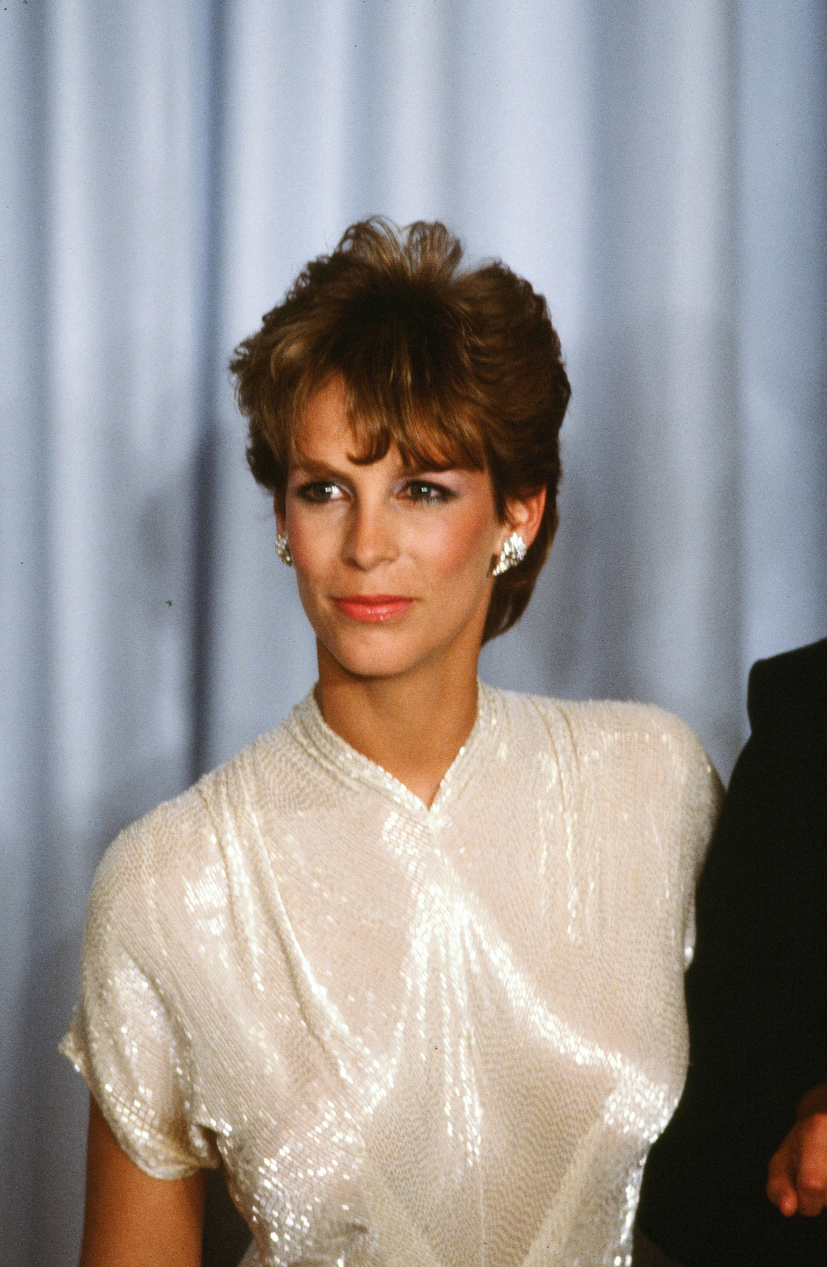 Jamie Lee Curtis radiates elegance backstage at the 55th Academy Awards, wearing a luminous ivory sequin gown with structured shoulders, complemented by sculpted hair, bold earrings, and a confident gaze.