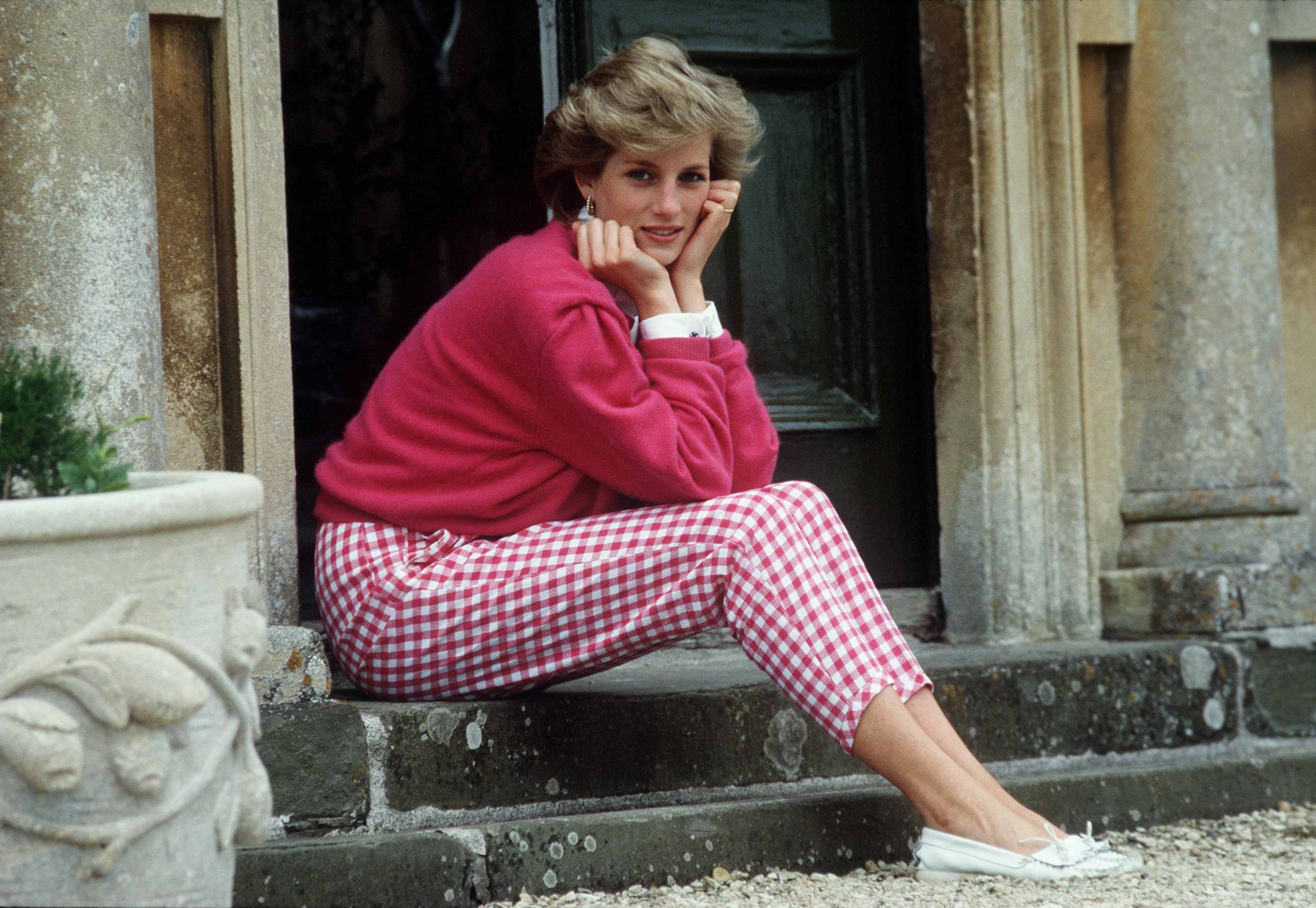 Photographed on July 18, 1986, Princess Diana is seen seated on the stone steps of Highgrove House in Gloucestershire — relaxed, radiant, and far from the formalities of royal life. Dressed in a vibrant pink jumper and red gingham trousers, she rests her chin on her hands with an easy smile.