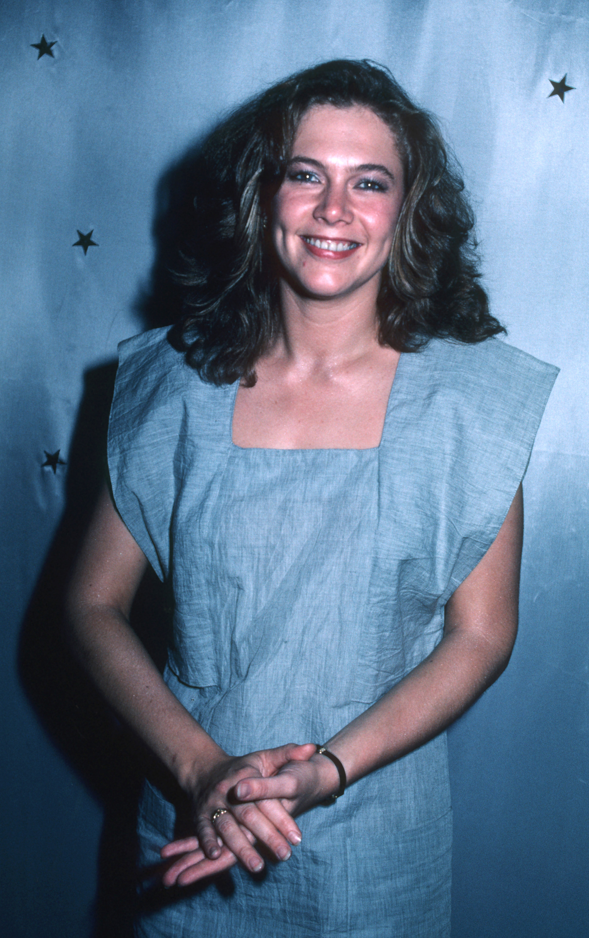 Actress Kathleen Turner is at the launch party for the new magazine "The Movies." She is smiling warmly while wearing a loose, sleeveless light blue dress with minimal accessories, embodying a relaxed yet stylish look.