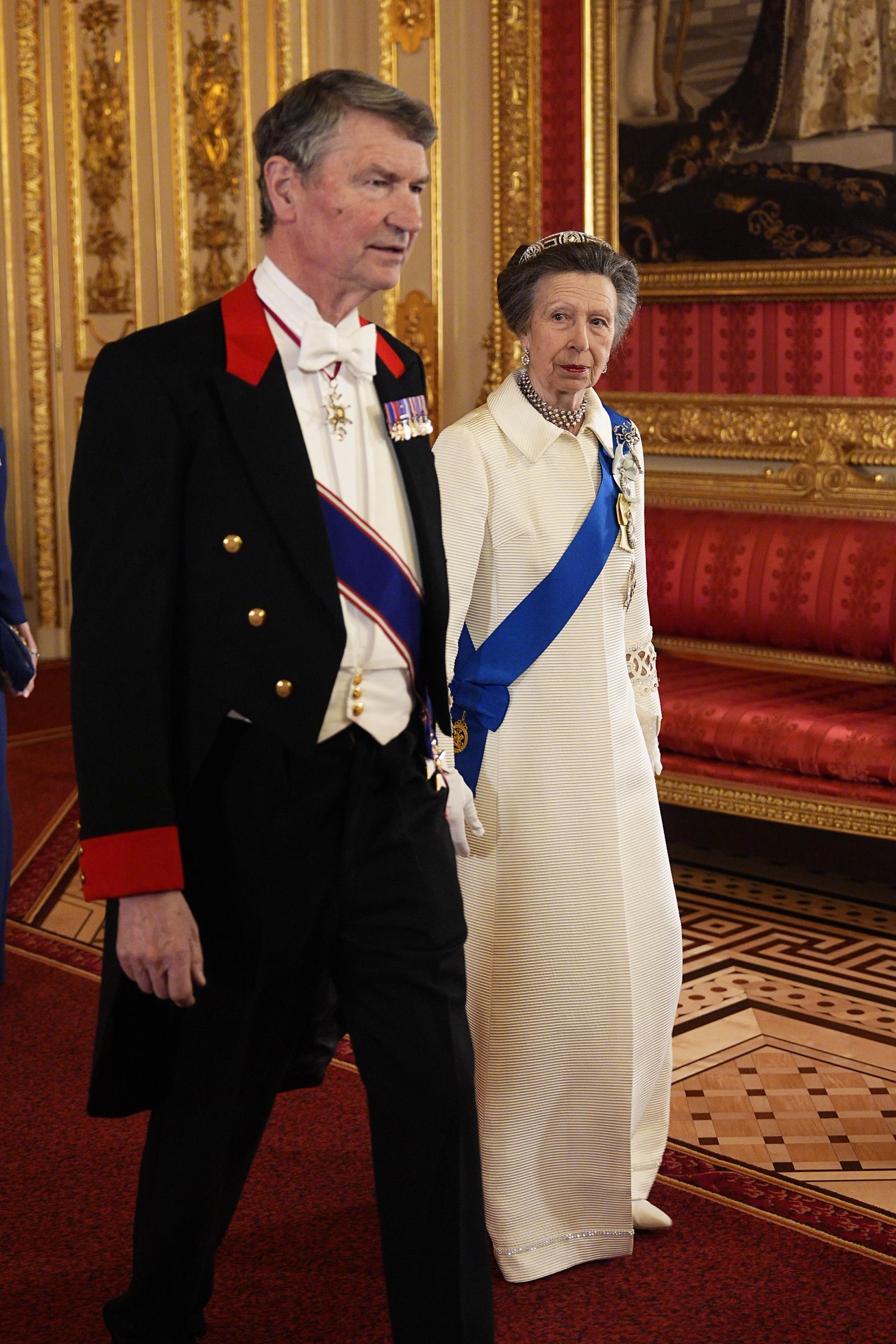 Princess Anne and Sir Timothy Laurence stand together in the gilded grandeur of Windsor Castle ahead of the state banquet on 18 March 2026. The coat's simple, plain collar — a contrast to its 1969 incarnation — is visible at Anne's neckline, while her blue sash, layered pearl necklace, and ceremonial honours complete the look.