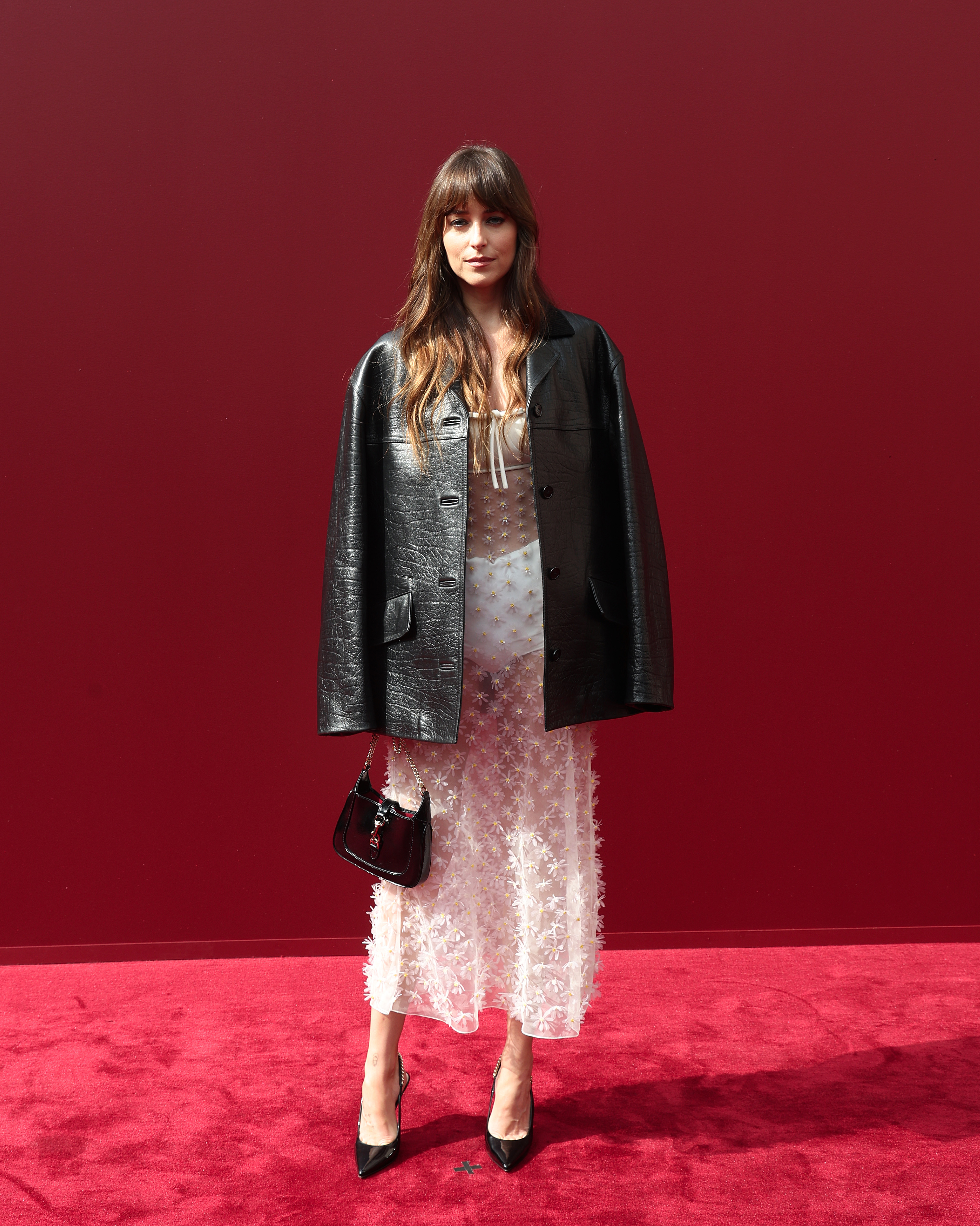 Dakota Johnson attends the Gucci Women's Spring/Summer Fashion Show looking effortlessly chic in a sheer white floral dress over a slip. Paired with an oversized black leather jacket, patent handbag, and soft makeup, she gives off a modern-meets-vintage look.