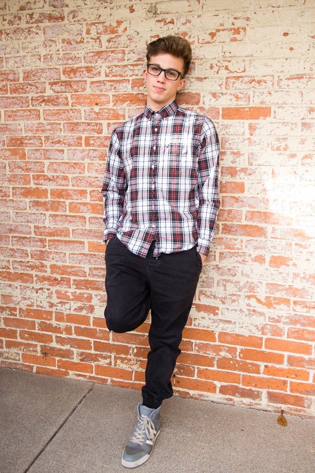 Boy wearing red and white plaid shirt and black pants leans against wall | Photo: Pexels