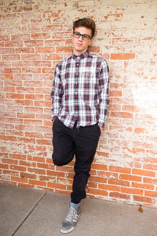 Young man wearing red and white plaid shirt and black pants leans against wall | Photo: Pexels