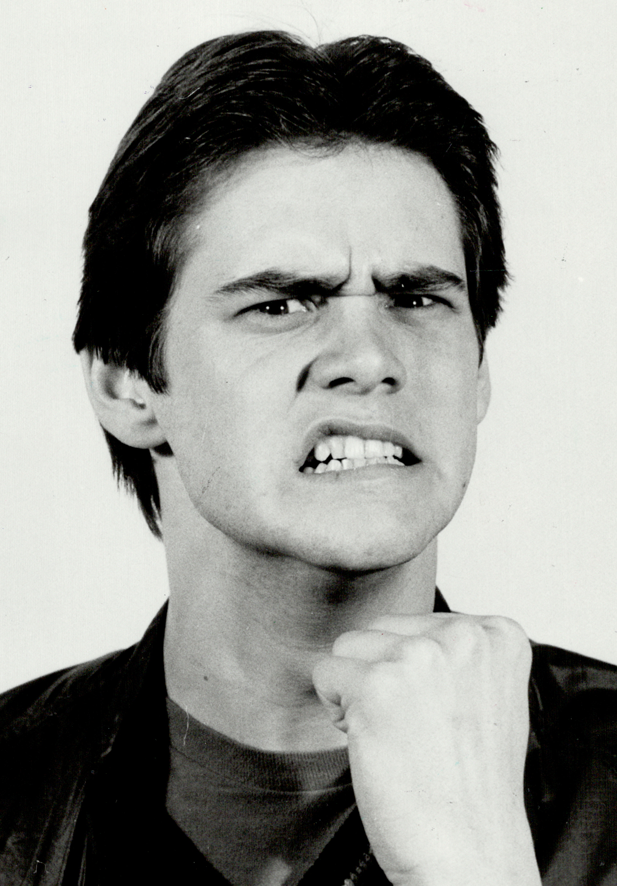 The black-and-white portrait shows a young Jim Carrey with a fierce, exaggerated expression, his brow furrowed and teeth clenched in mock determination. His fist is raised near his chin, enhancing the dramatic and comedic intensity of the pose. The image perfectly captures his signature blend of humor and expressive physicality.