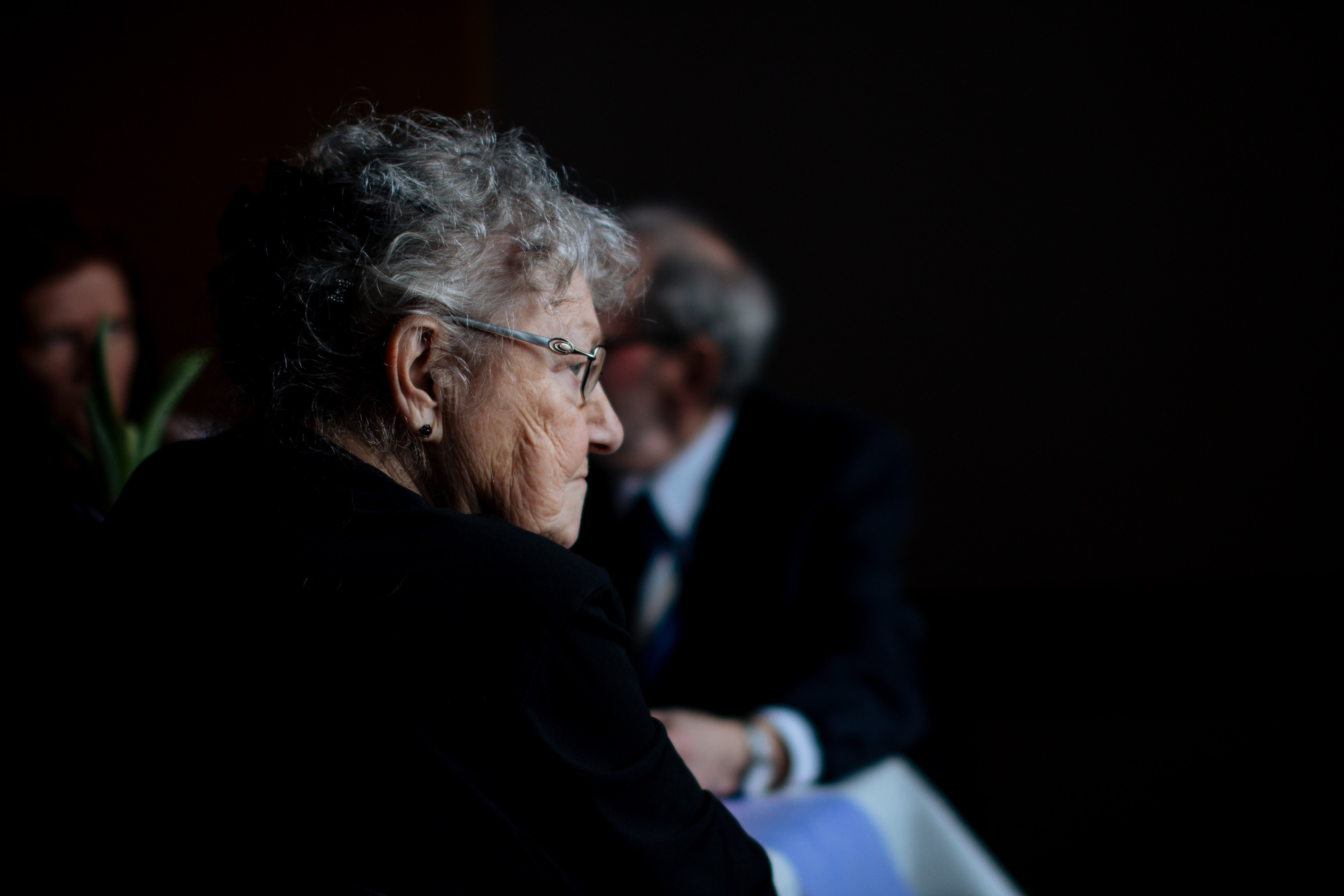 An older woman sitting by herself looking around while others appear in the background | Sources: Unsplash