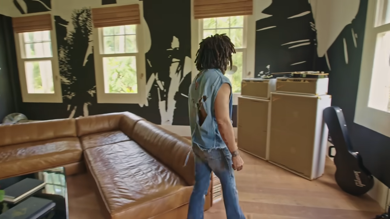 Lenny walks past a large tan leather sectional sofa in the bedroom lounge, with stacked Fender amplifiers and a guitar case visible in the corner. | Source: YouTube/@Archdigest