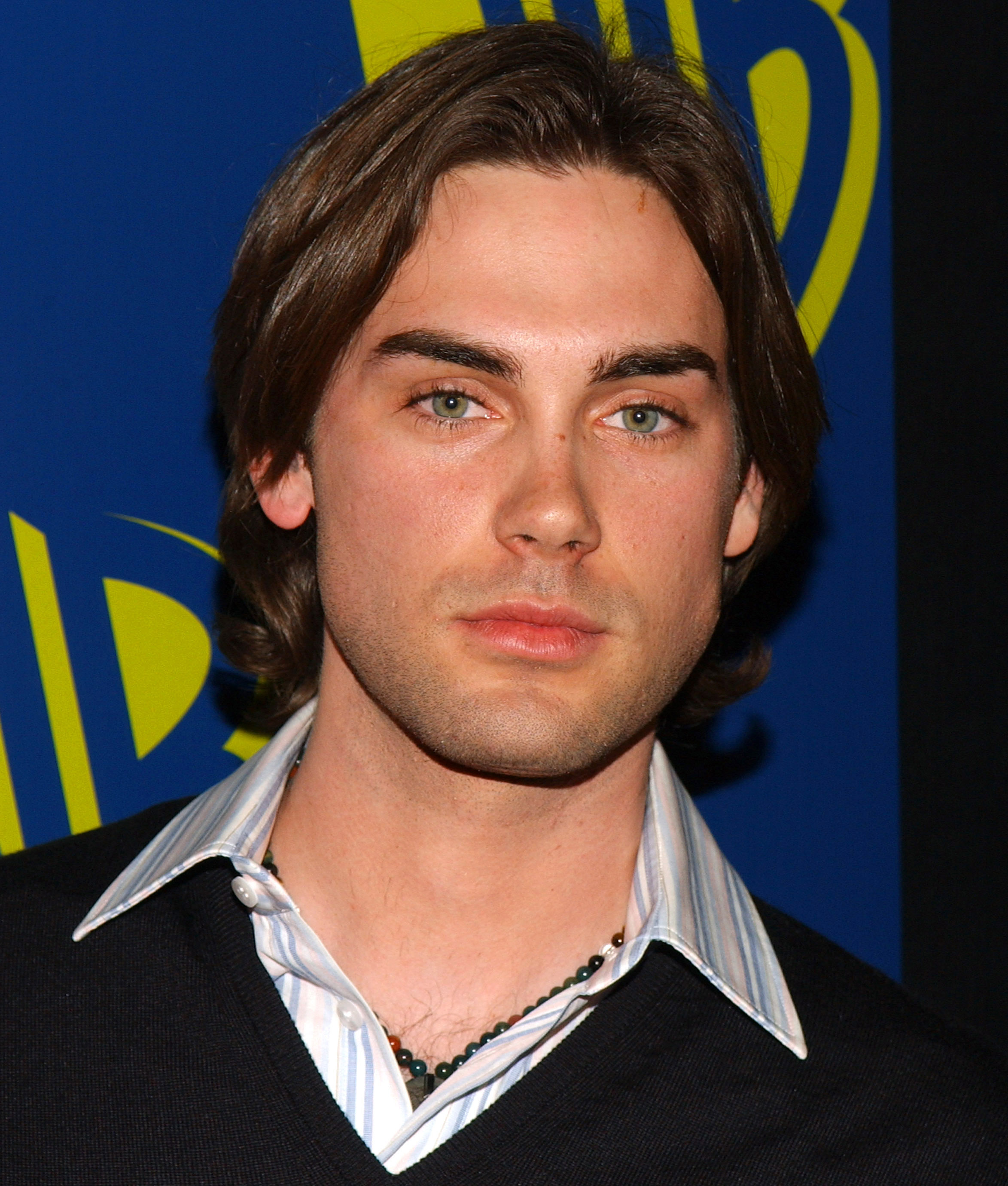 Drew Fuller is seen at a WB network event in the mid-2000s, wearing a striped collared shirt layered under a dark V-neck sweater. His shoulder-length hair and serious expression give the portrait a polished, youthful intensity, fitting for his role during his time on "Charmed".