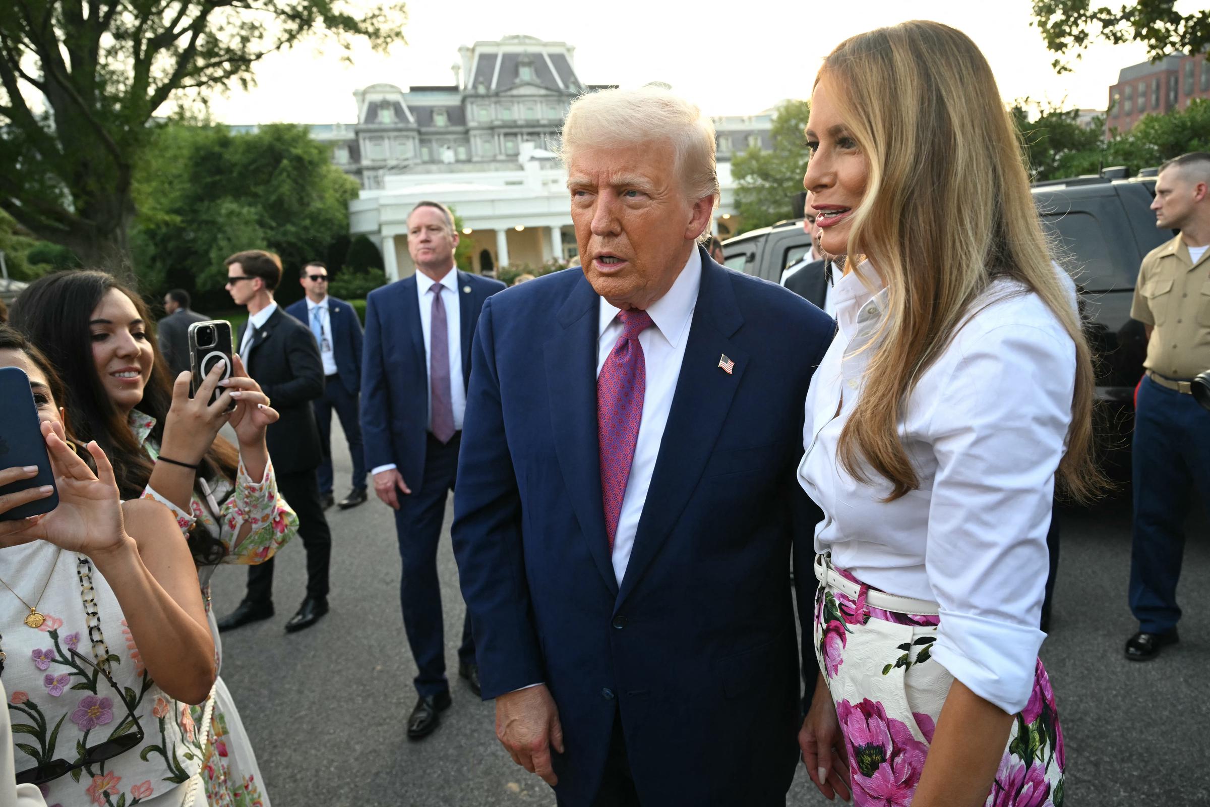 Donald and Melania Trump greet more guests near the White House driveway, surrounded by smiling attendees snapping photos. The background reveals security detail and historic buildings, grounding the event in its iconic location.