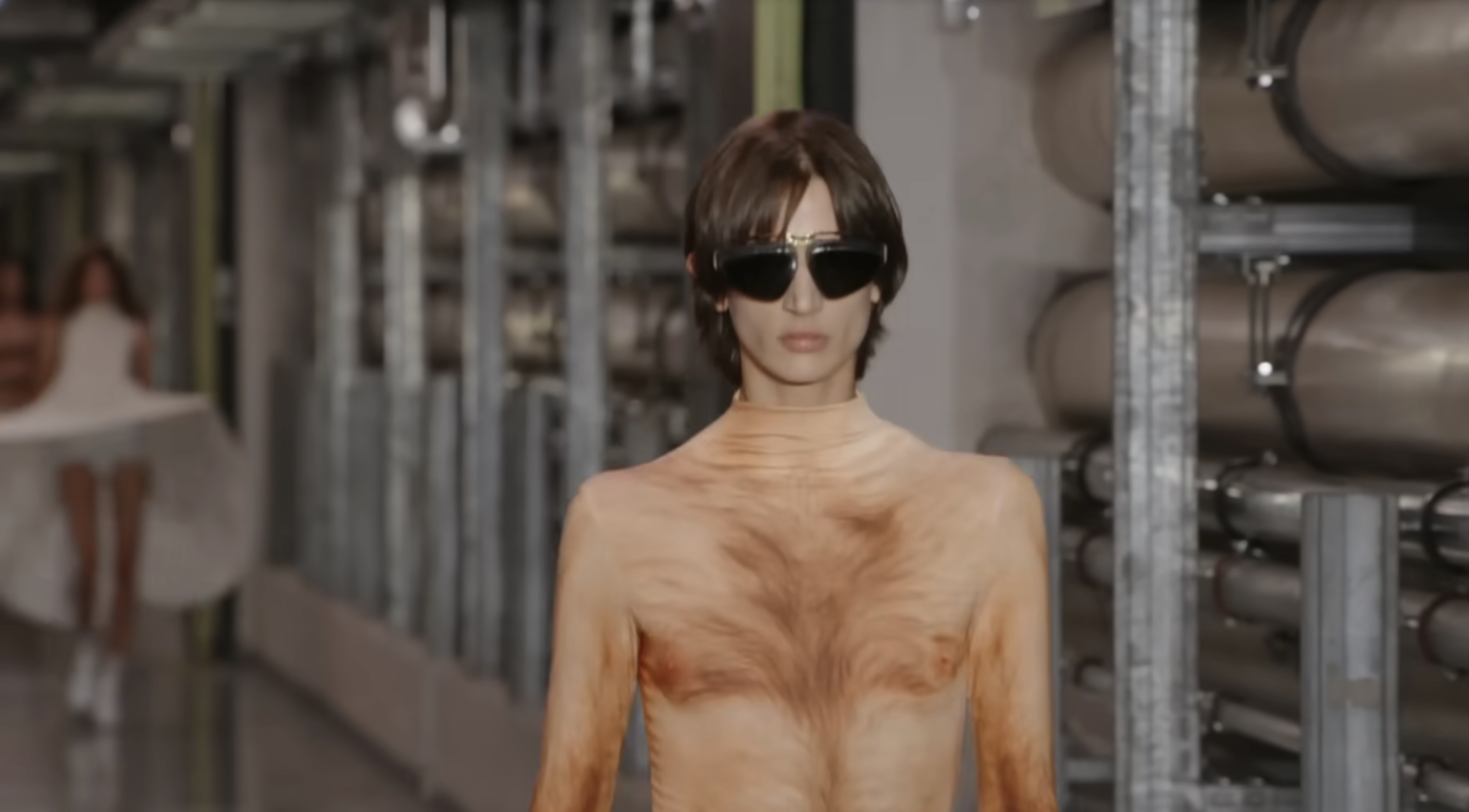However, a closer camera shot revealed that the model was actually wearing a jumpsuit beneath the skirt. The piece is nearly indistinguishable at first glance, matching his skin tone and featuring a printed body-hair design that creates the illusion of bare skin. From a post dated October 5, 2025. Source: Youtube/@jeanpaulgaultier