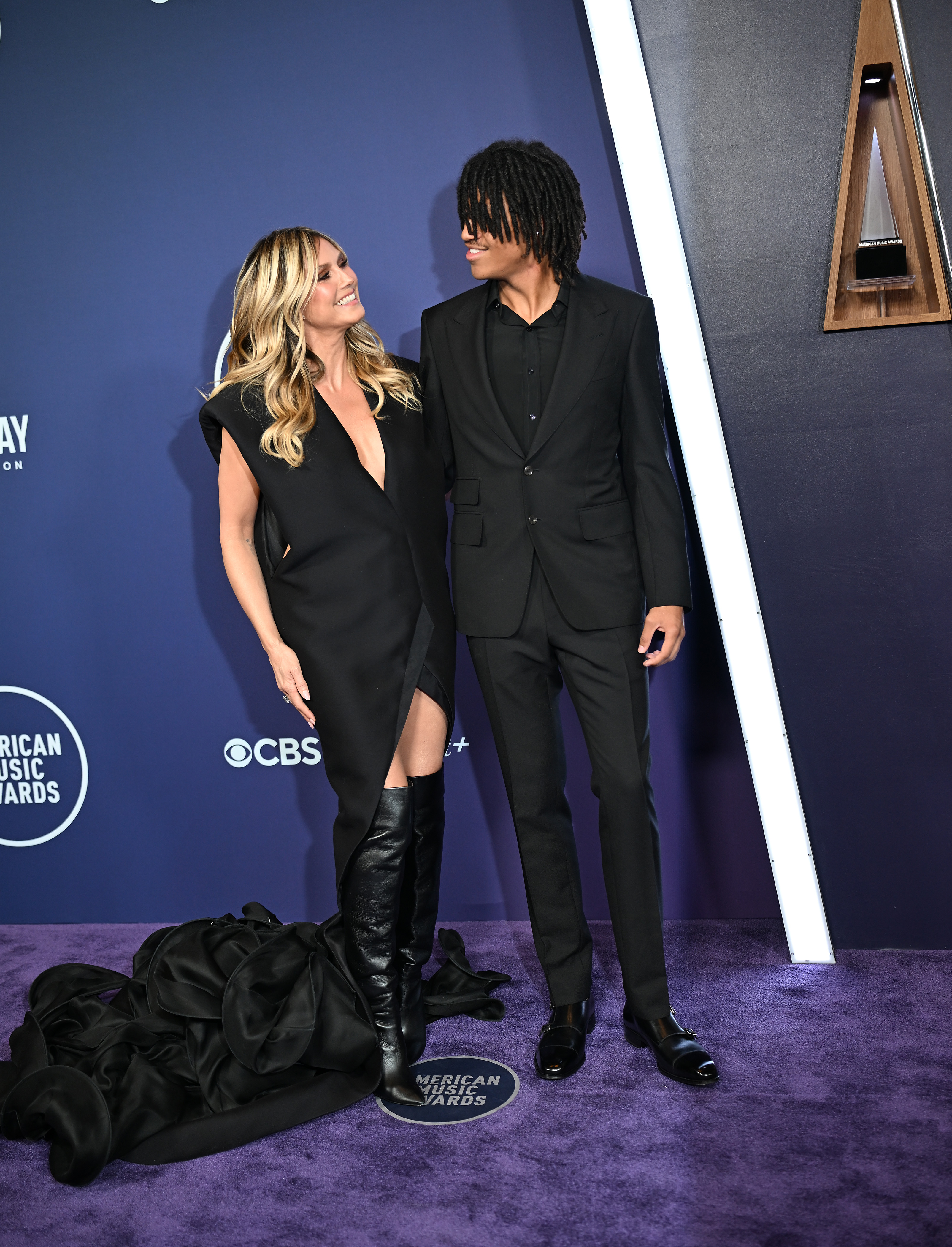 Heidi Klum shares a warm moment with her son, Henry Samuel, as they exchange smiles on the AMAs red carpet — her dramatic train pooled beside his sharp black suit.