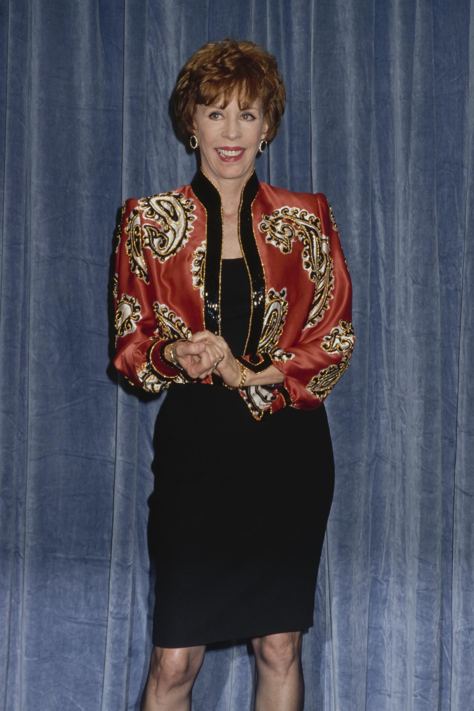 Posing in front of a curtain backdrop, she wears a black dress topped with a structured red jacket featuring ornate gold detailing. Her hands are clasped together in front of her.