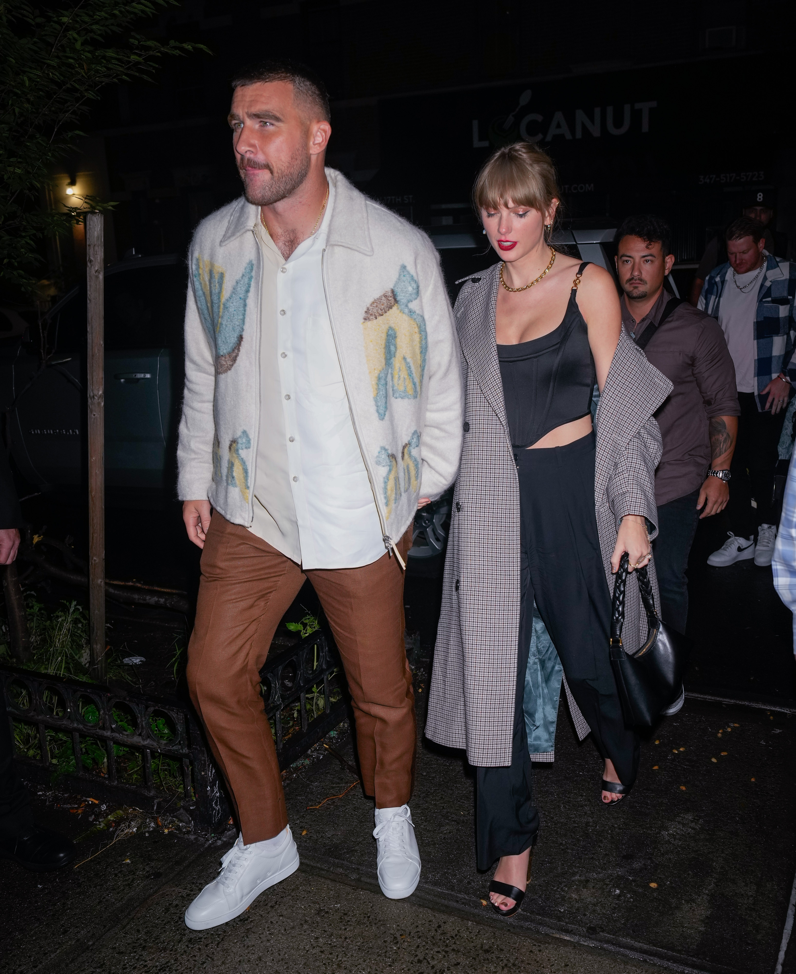 Travis Kelce, wearing a white shirt layered under a patterned cream jacket with brown pants and white sneakers, walks hand in hand with Taylor Swift, who is dressed in a black top with cutouts, black pants, and a long gray checkered coat, as they arrive at the "Saturday Night Live" afterparty on October 15, 2023, in New York City.