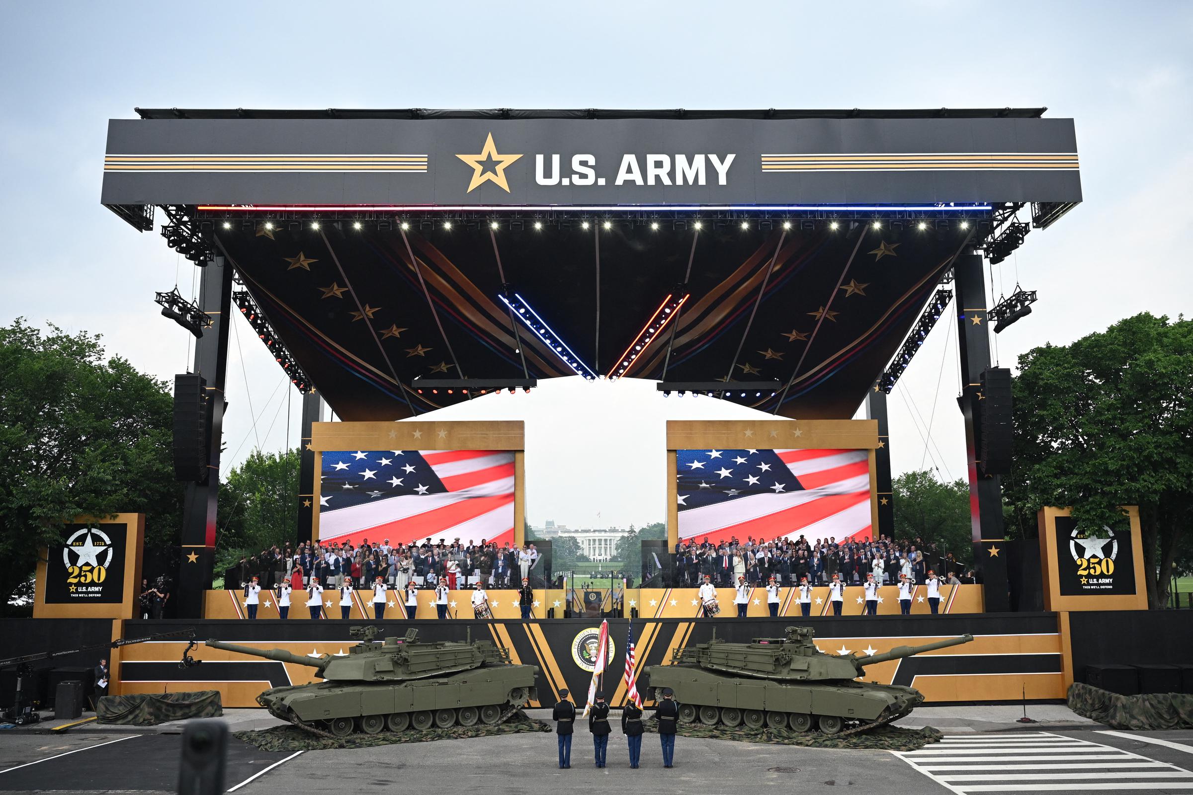 The grand stage at the US Army's 250th anniversary parade features American flags, tanks, and uniformed personnel. The event is framed by a massive U.S. Army banner, with choreographed formations and vibrant lighting in full view.