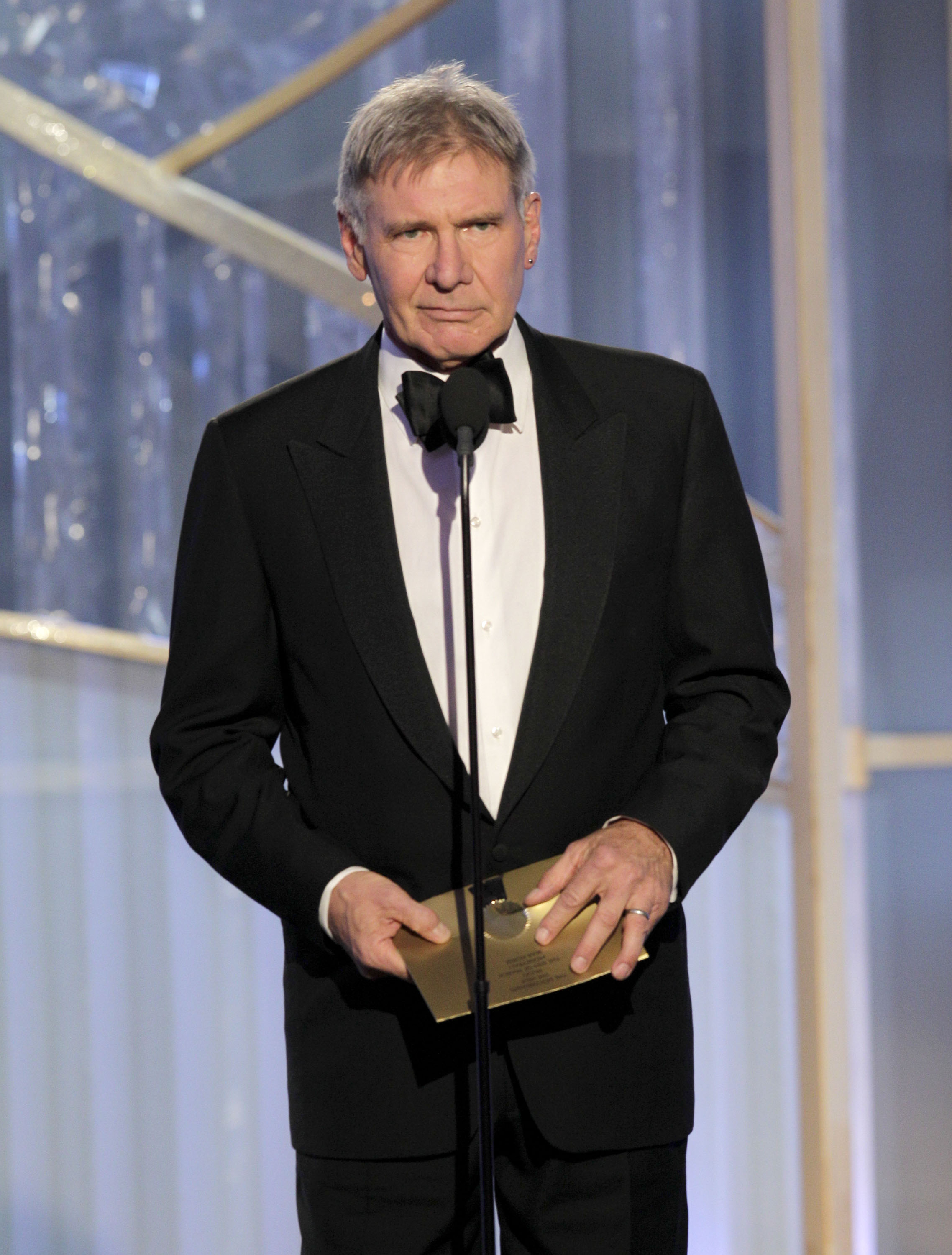 Harrison Ford takes the stage at the Beverly Hilton Hotel as a presenter during the 69th Annual Golden Globe Awards. Clad in a traditional black tuxedo and matching bow tie, the actor holds a gold envelope at the microphone. | Source: Getty Images