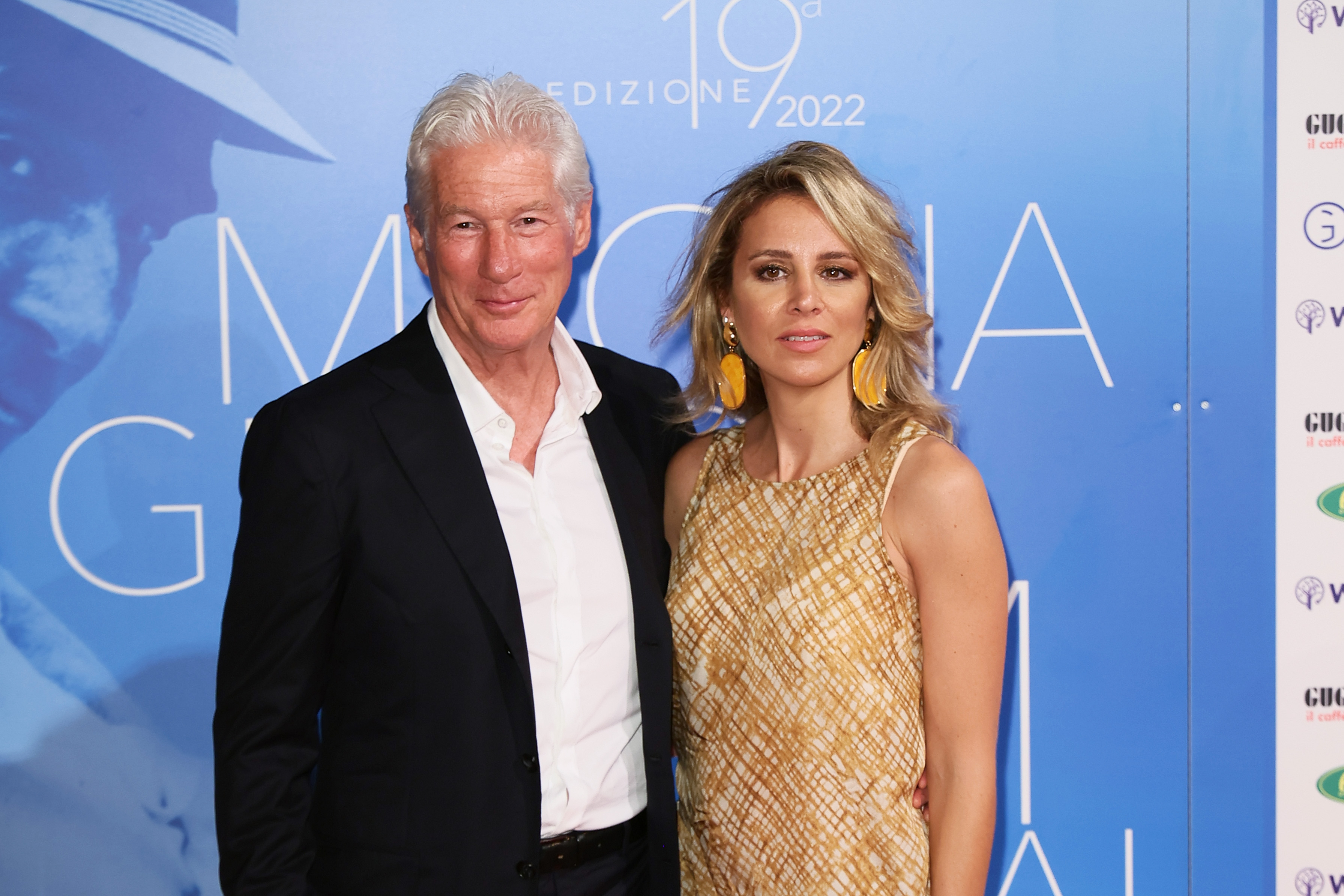 At the Magna Graecia Film Festival in Catanzaro on August 5, 2022, Richard Gere brought his signature charm in a crisp black suit, while Alejandra Silva lit up the red carpet in a golden patterned dress and bold earrings, effortlessly stealing the spotlight.