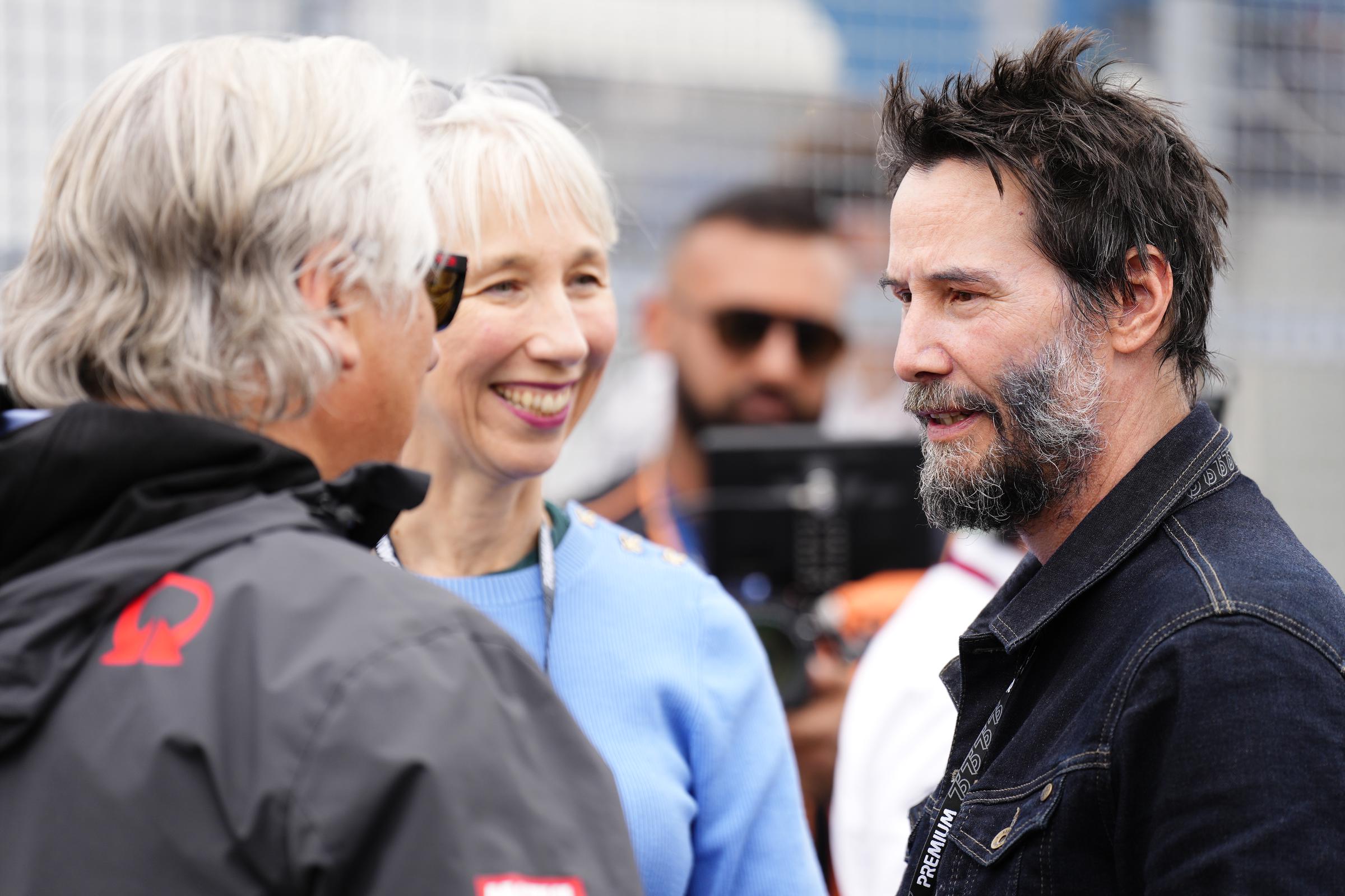 Alexandra Grant smiles as she looks at Keanu Reeves talking to a guest at the Liqui Moly Motorrad Grand Prix Deutschland at Sachsenring Circuit in Hohenstein-Ernstthal, Germany on July 7, 2024. | Source: Getty Images