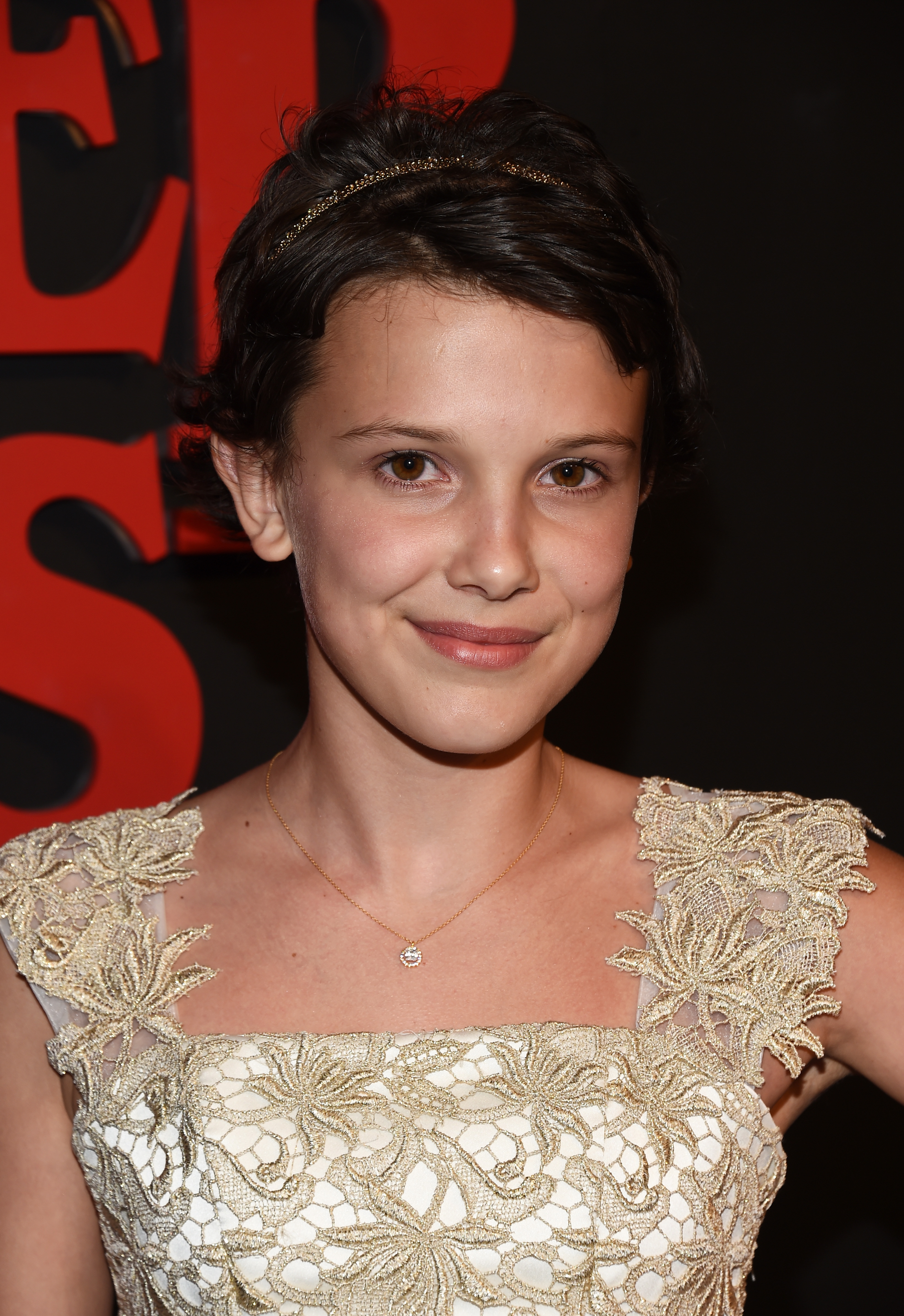With softly cropped hair and minimal makeup, Millie Bobby Brown appears fresh and quietly radiant. Delicate gold embroidery frames her neckline, lending the look a timeless elegance. Her gentle smile adds warmth to an otherwise polished, classic appearance.