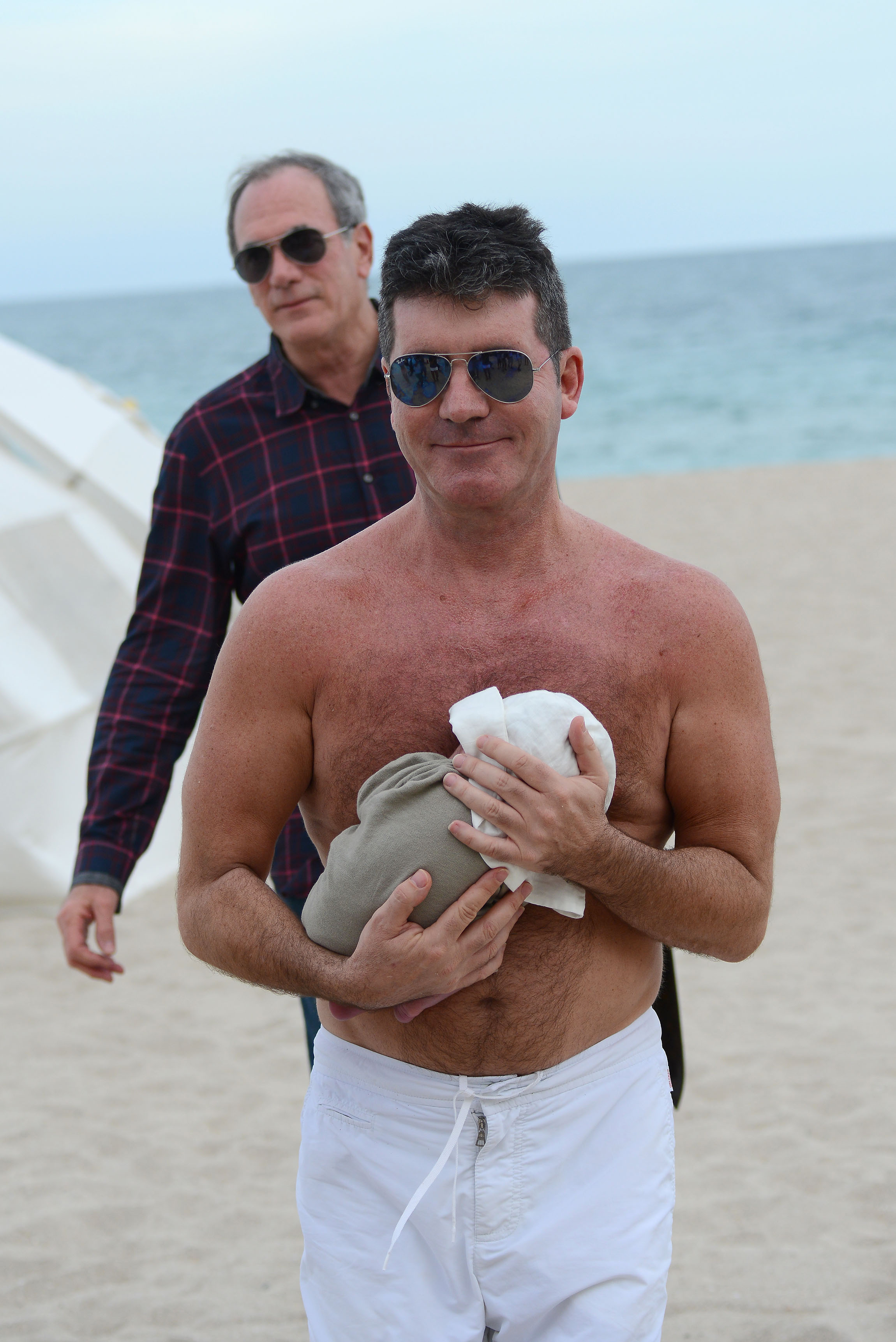 Simon Cowell, joined by a friend standing behind him, is pictured at the beach in Miami, Florida, cradling his newborn son Eric against his bare chest. The tiny baby rests snuggly on him as Cowell smiles at the camera.