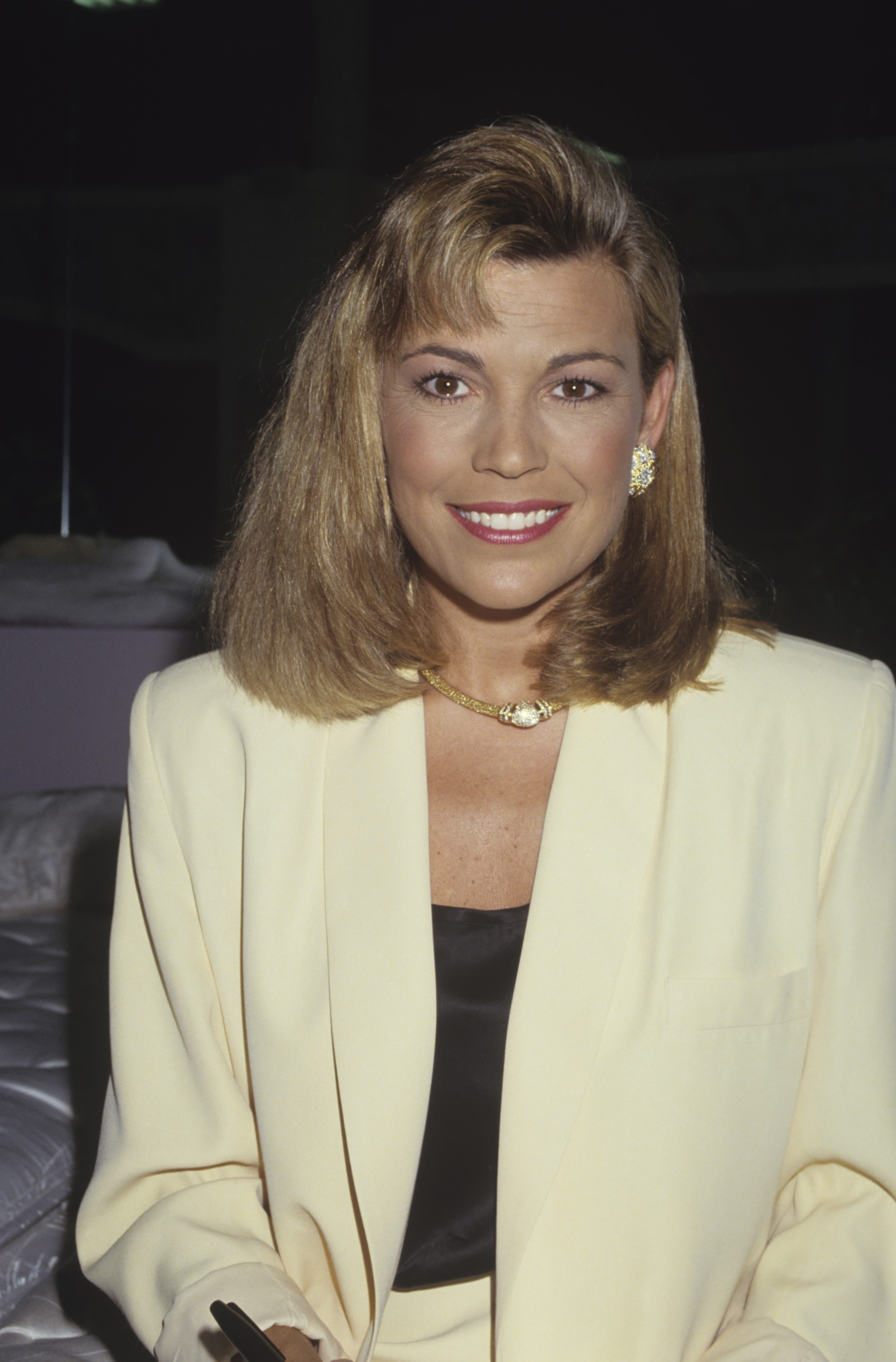 Vanna White smiles warmly in a cream blazer over a dark top, accessorized with gold jewelry, in a portrait highlighting her fame as the hostess and puzzle-board turner of "Wheel of Fortune."