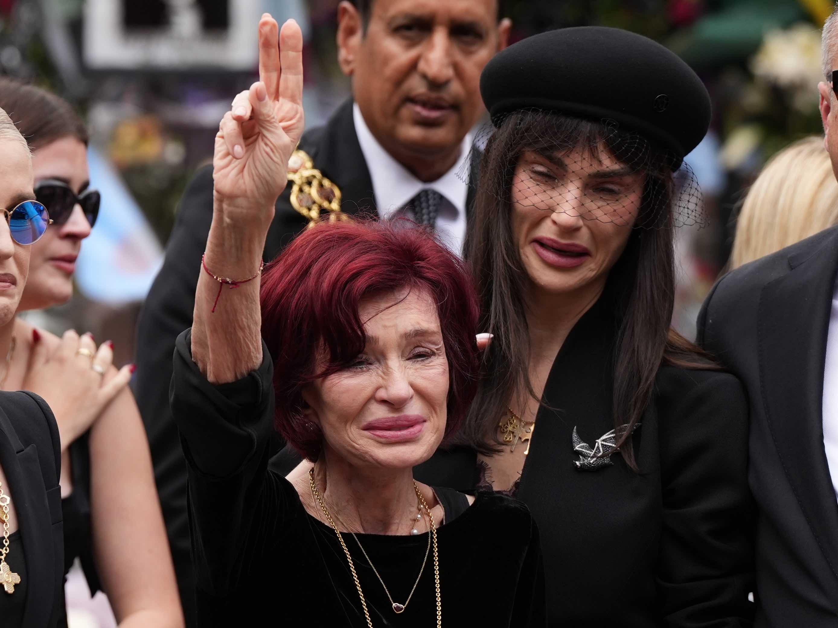Her face etched with sorrow, Sharon Osbourne holds up a tearful peace sign at her husband's funeral. Their daughter, Aimee is also distraught and overcome with grief during the funeral procession.