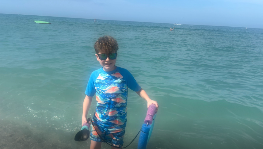 One of Amanda "Mandy" Karolkiewicz and Kristopher Karolkiewicz's sons stands in shallow water holding pool noodles while wearing a blue rash guard and sunglasses during the family's spring break vacation. | Source: TikTok/mandykarolk