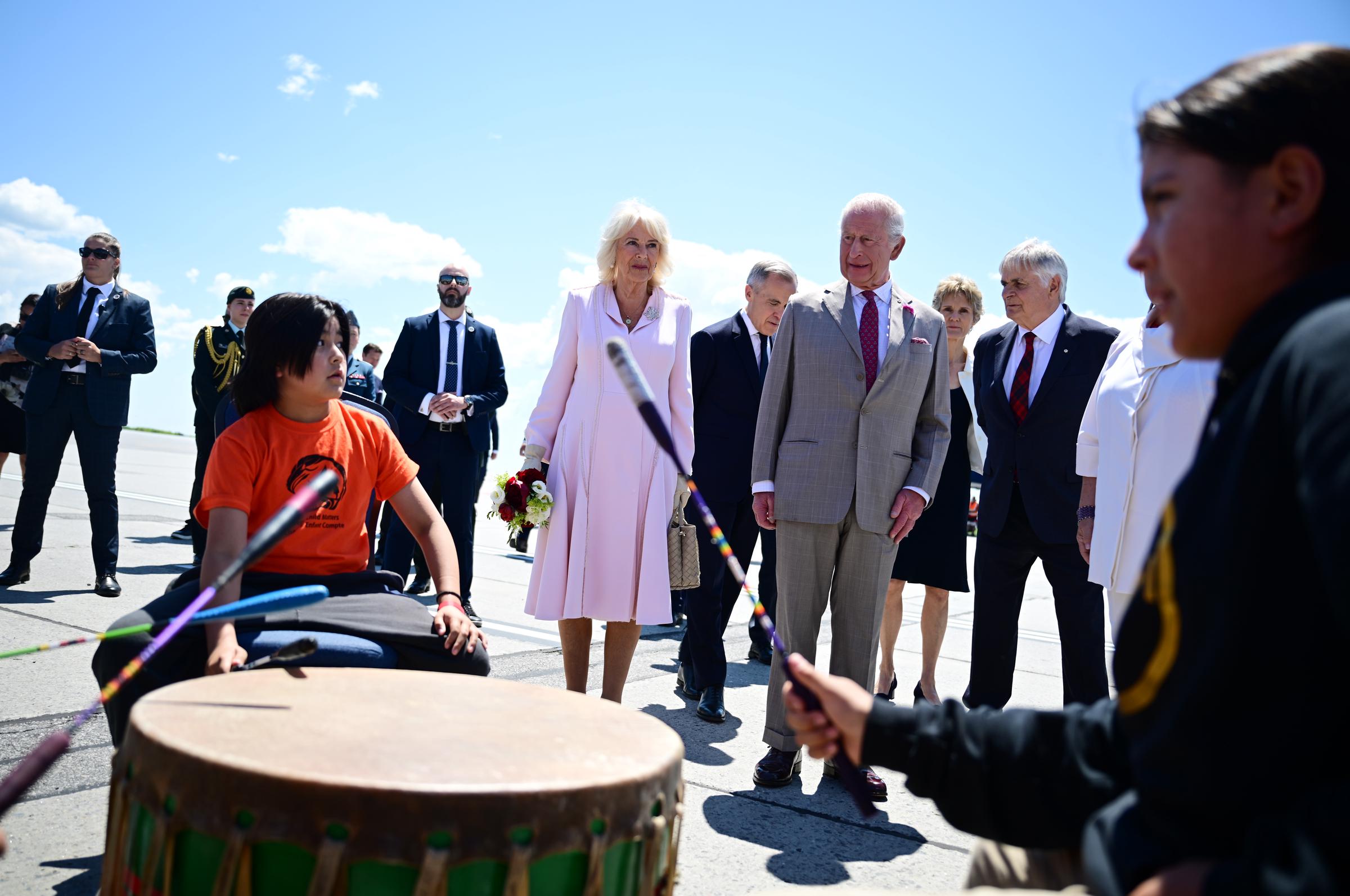 A vibrant display of pomp and pageantry greets King Charles III and Queen Camilla as they step onto Canadian soil, with artists gathered around, rhythmically beating drums in a spirited welcome.