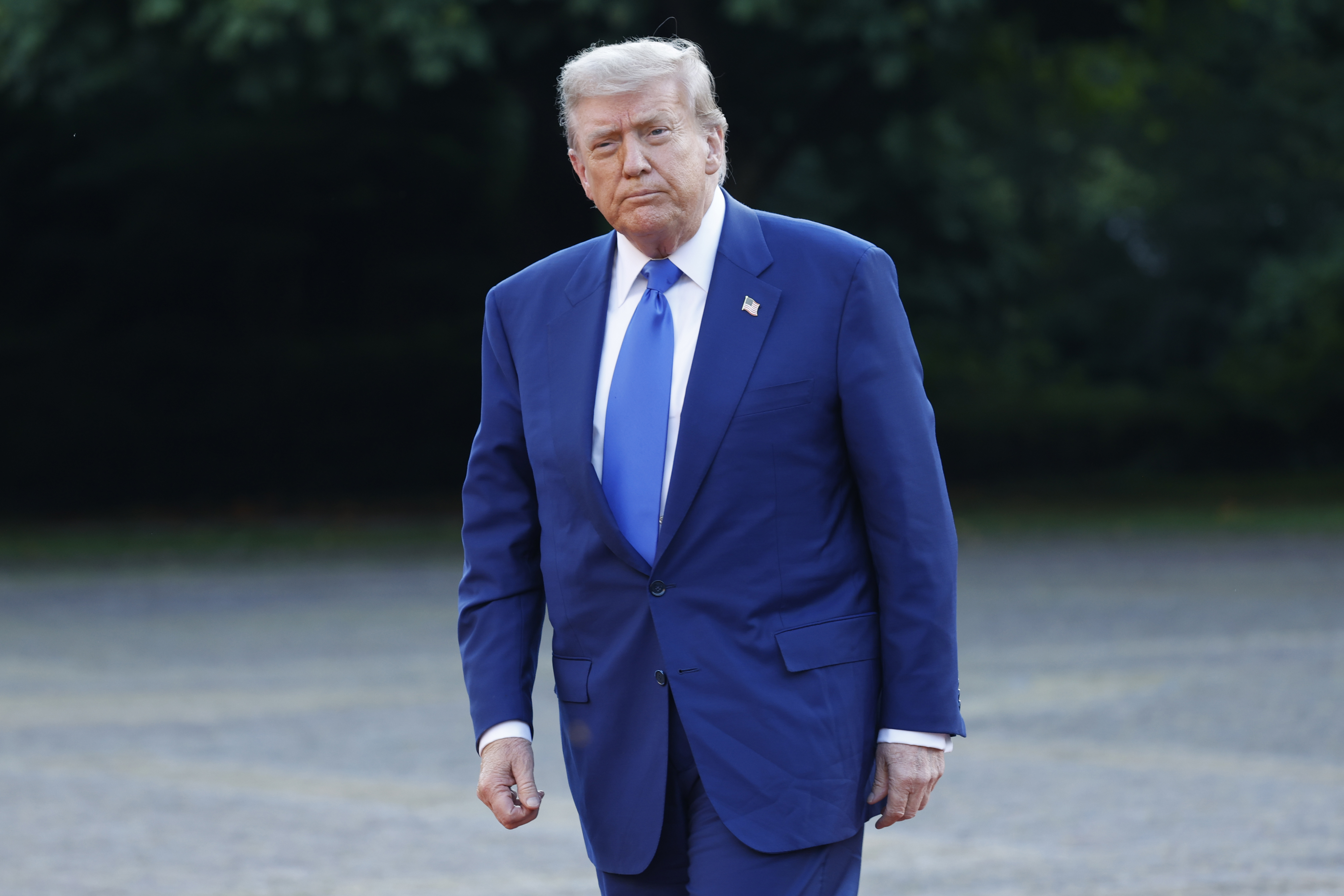 Trump wears a serious, presidential expression as he walks in a sharp blue suit paired with a slightly lighter-shaded blue tie. A small U.S. flag lapel pin stands out on his jacket, adding a patriotic touch to his formal appearance during the high-level NATO gathering.