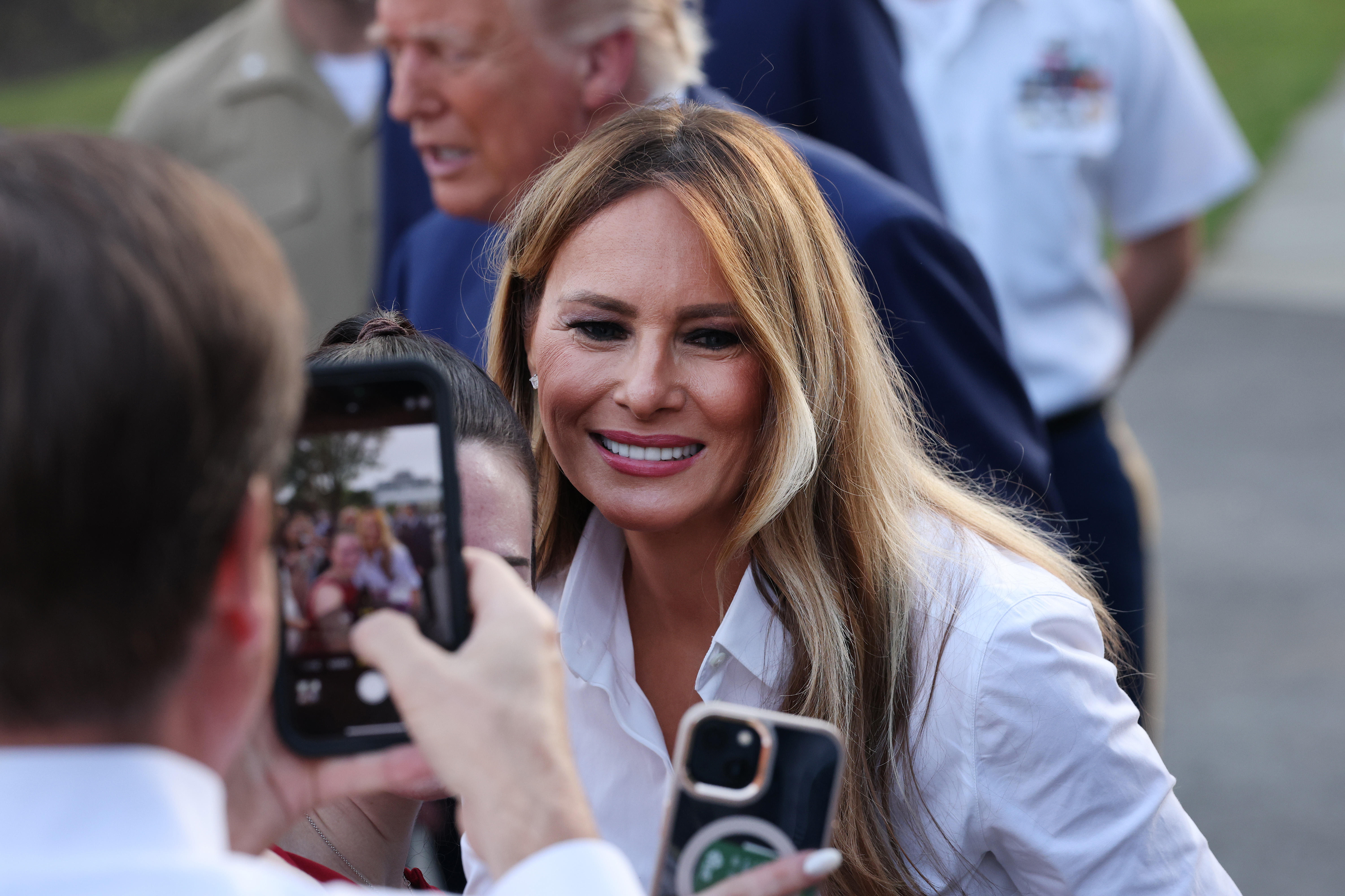 Melania Trump smiles warmly while posing for a photo with attendees during the 2025 Congressional Picnic. Surrounded by guests and phones capturing the moment, she engages directly with the crowd in a relaxed, approachable setting.