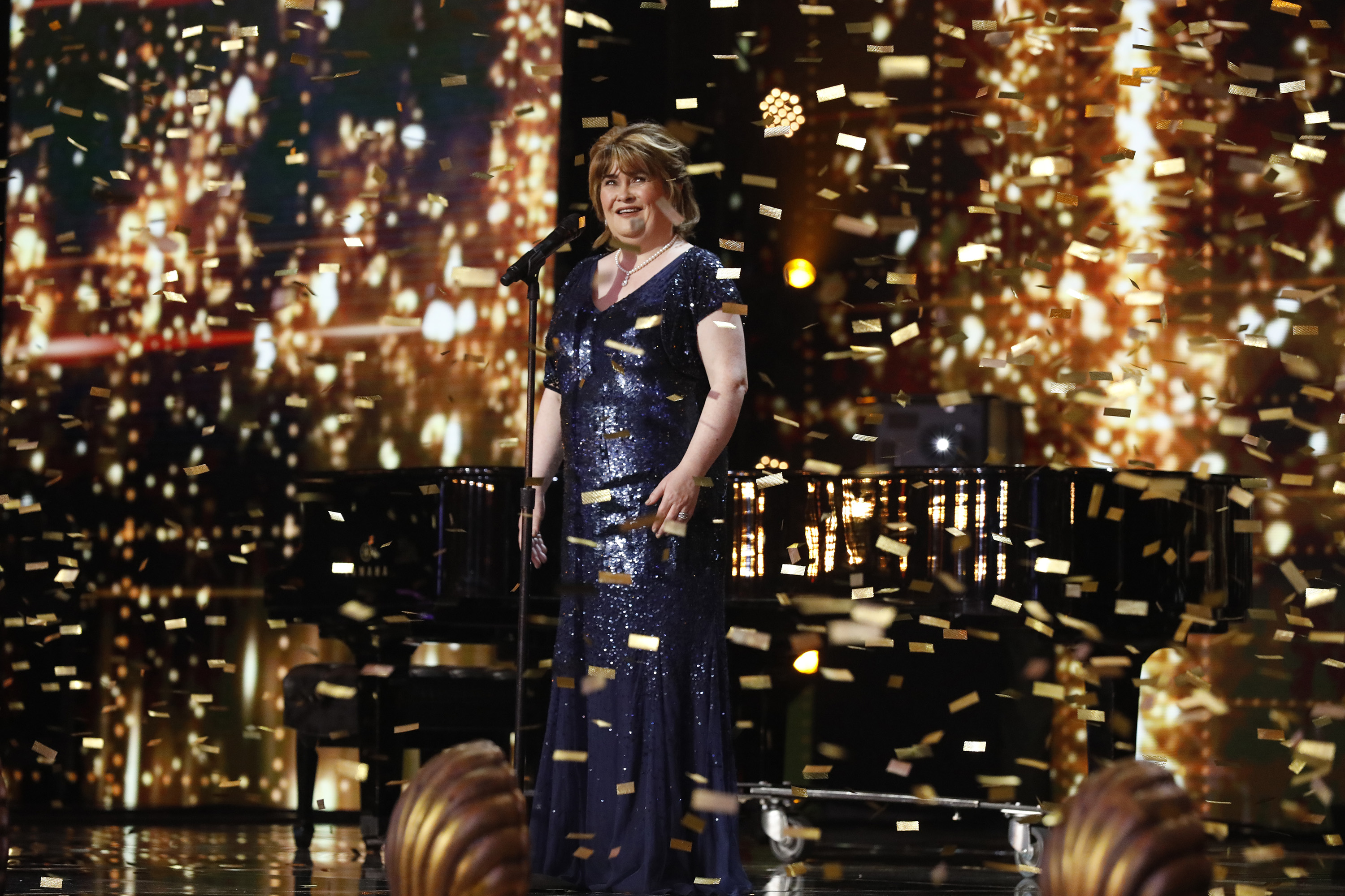 Boyle stands in stunned delight at a microphone stand as a cascade of gold confetti rains down around her during "America's Got Talent: The Champions," wearing a floor-length navy sequinned gown.