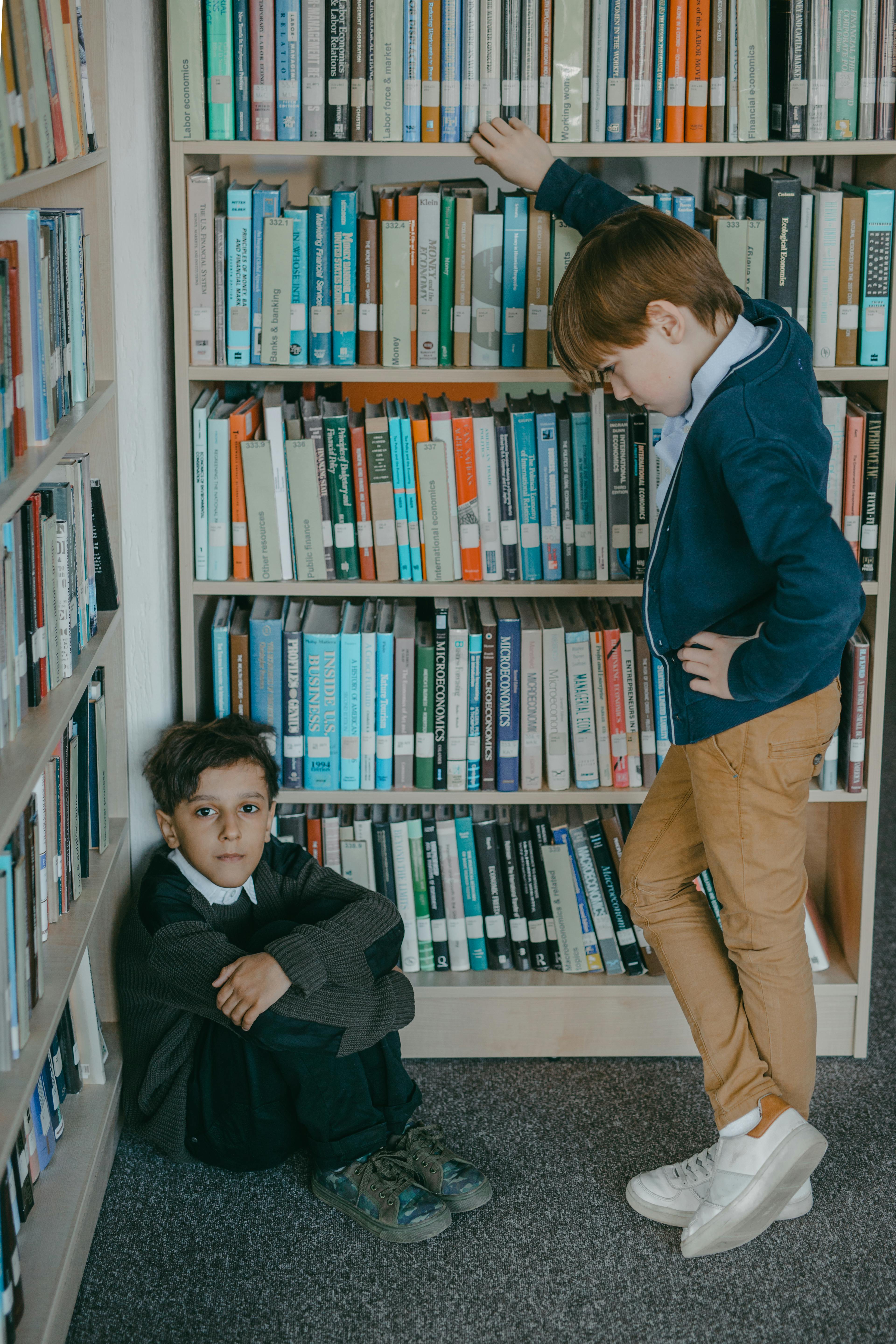 A young boy sits alone beside a bookshelf while another child stands over him, highlighting the imbalance of power often seen in bullying situations. | Source: Pexels