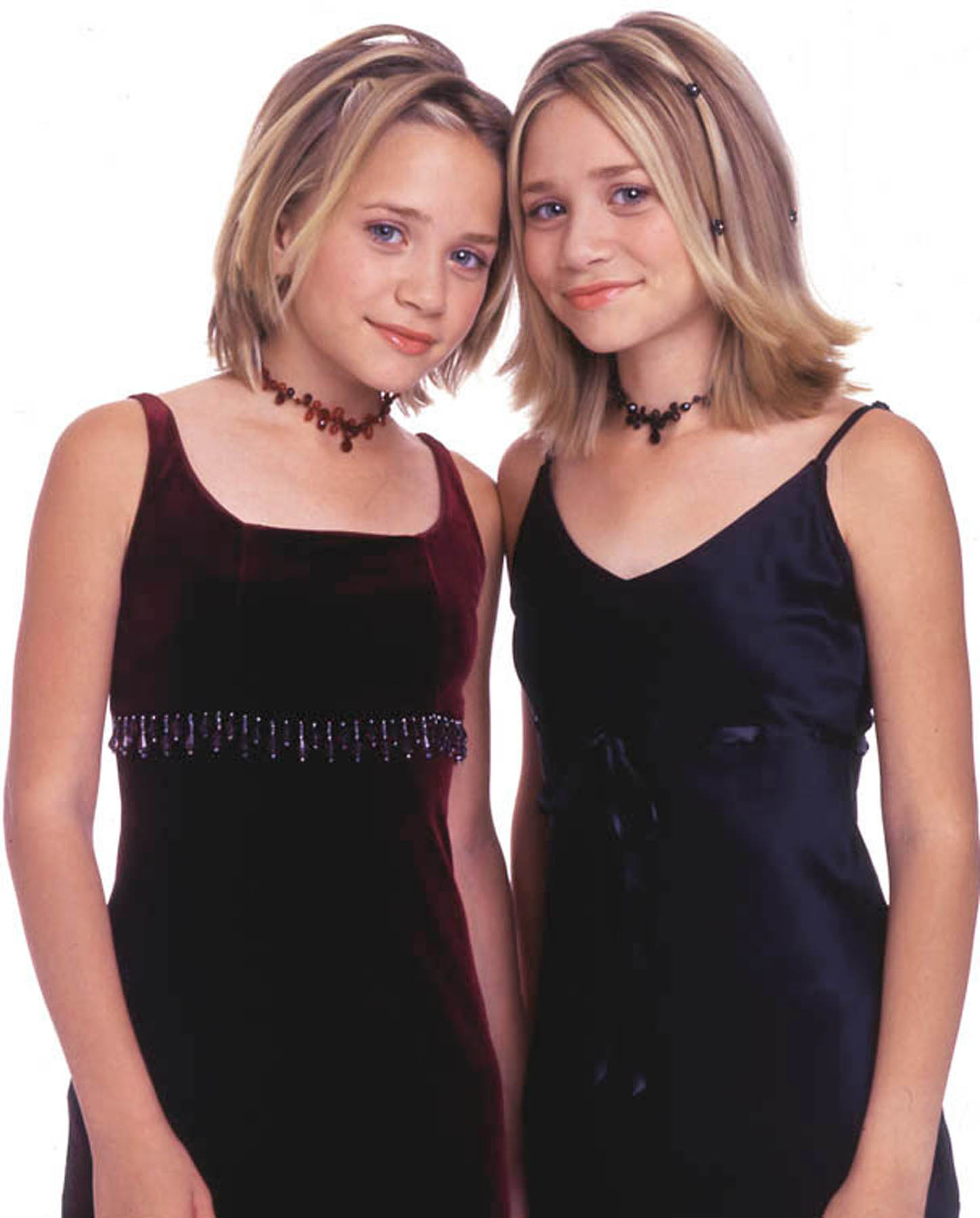 Mary-Kate Olsen and Ashley Olsen stand shoulder to shoulder in sleeveless dresses and chokers. They angle inward with soft smiles and a polished, composed look.