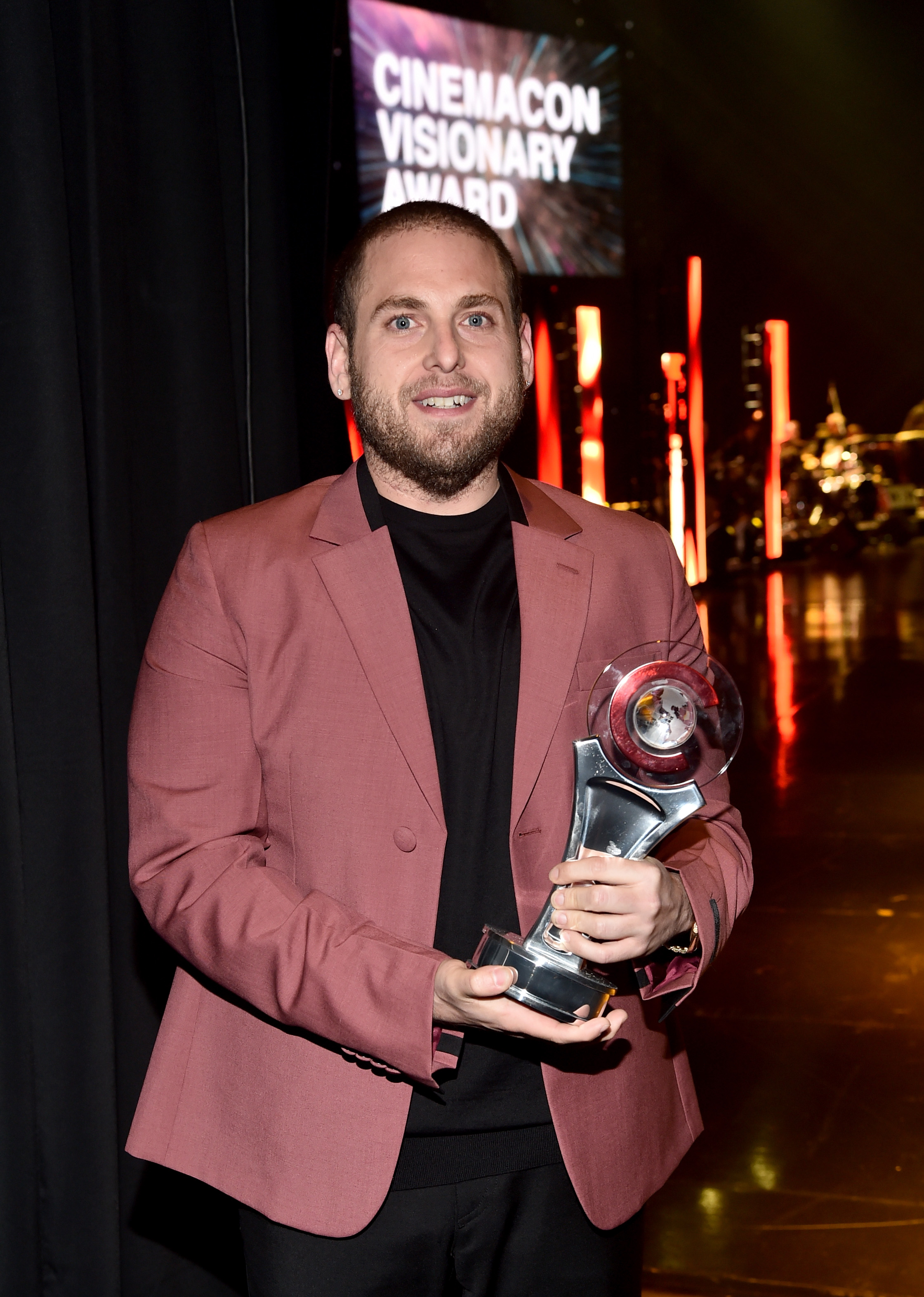 Jonah Hill poses backstage with his CinemaCon Visionary Award, wearing a dusty rose suit jacket over a black shirt. Smiling proudly, he holds the trophy with both hands as the stage glows behind him.