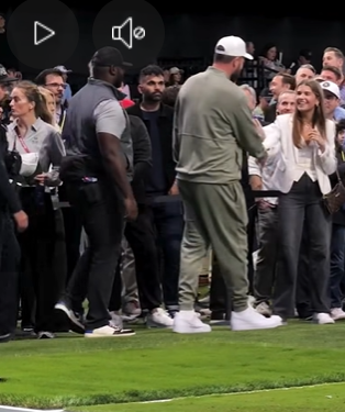 Kai Trump smiles as Travis Kelce approaches and shakes her hand while security and spectators gather nearby at the edge of the turf field. | Source: Facebook/PageSix